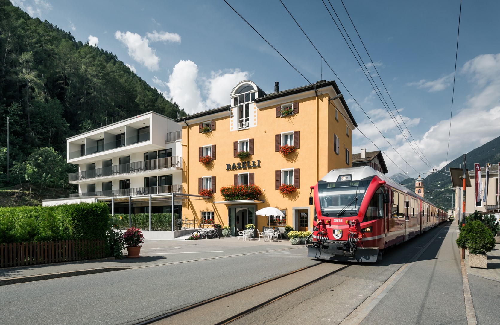 A red RhB train standing at the station in Le Prese. Right next door, the Raselli Sport Hotel.