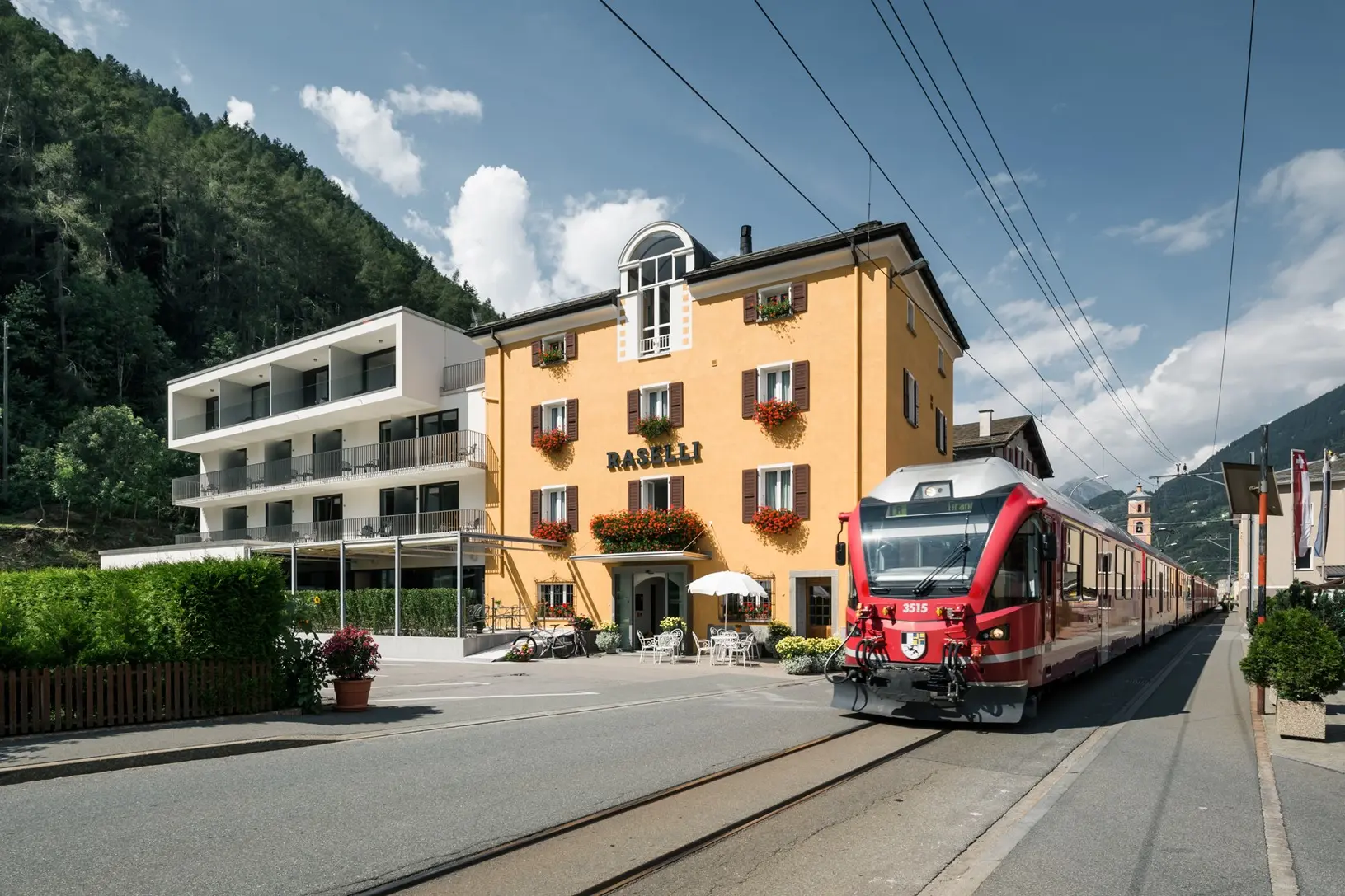 A red RhB train standing at the station in Le Prese. Right next door, the Raselli Sport Hotel.
