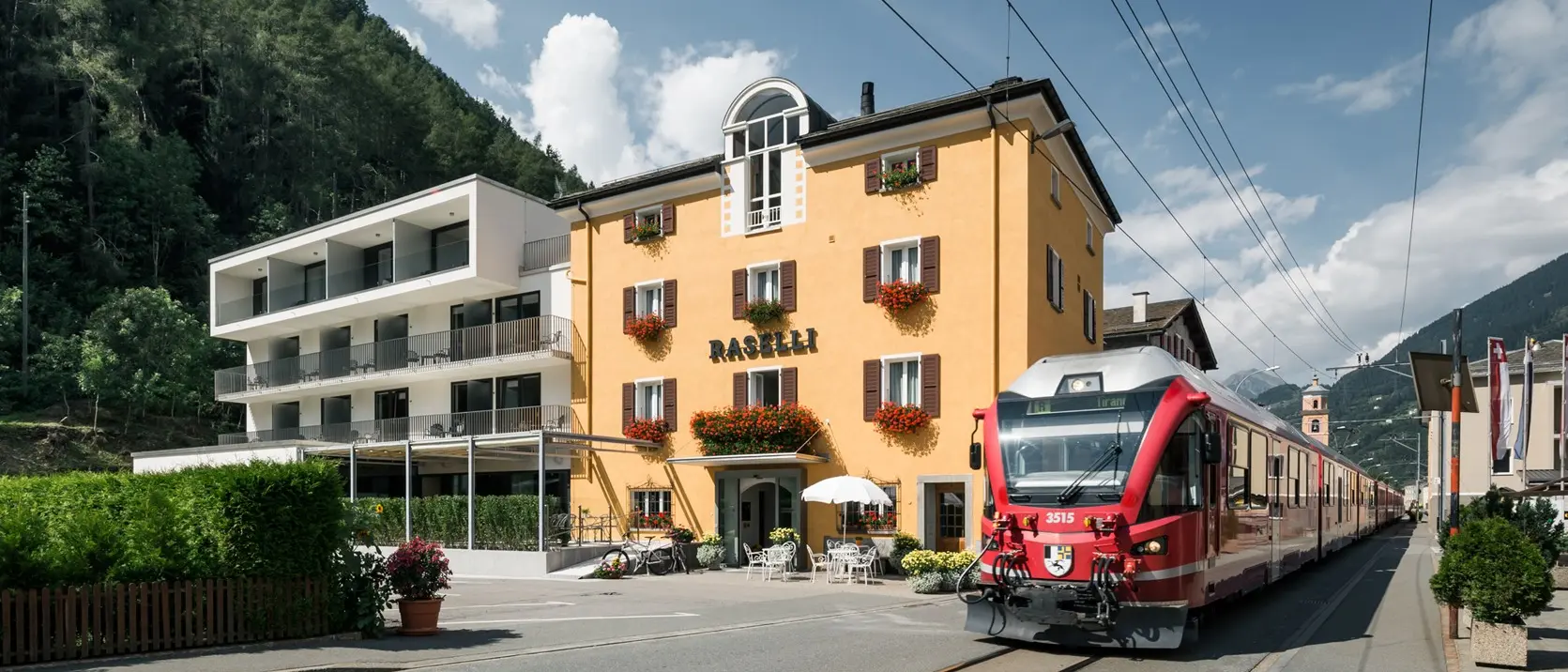 A red RhB train standing at the station in Le Prese. Right next door, the Raselli Sport Hotel.