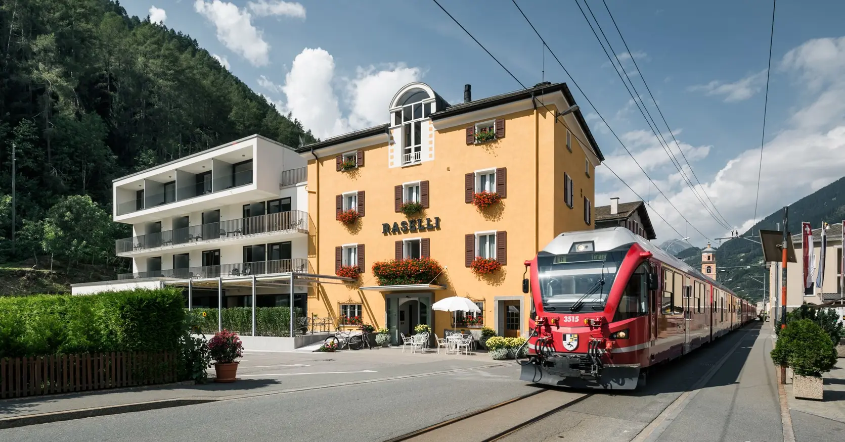 A red RhB train standing at the station in Le Prese. Right next door, the Raselli Sport Hotel.