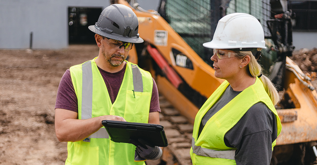 A construction worker wearing a high-visibility lime green safety vest, a dark hard hat, safety glasses, and holding a tablet.