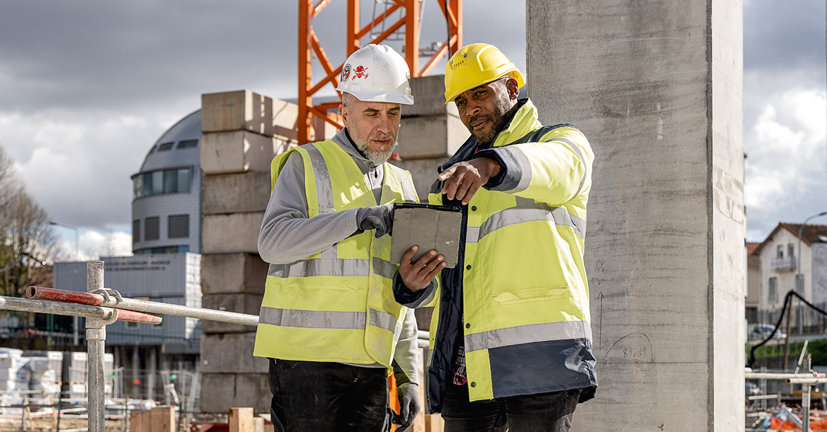 Two construction workers in high-visibility jackets and hard hats reviewing plans on a tablet at an active building site, illustrating the use of construction management software for on-site coordination and project tracking.