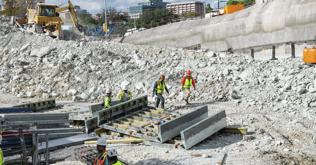 Construction workers in safety vests and helmets working on a large excavation site with rubble piles, concrete panels, and heavy machinery.