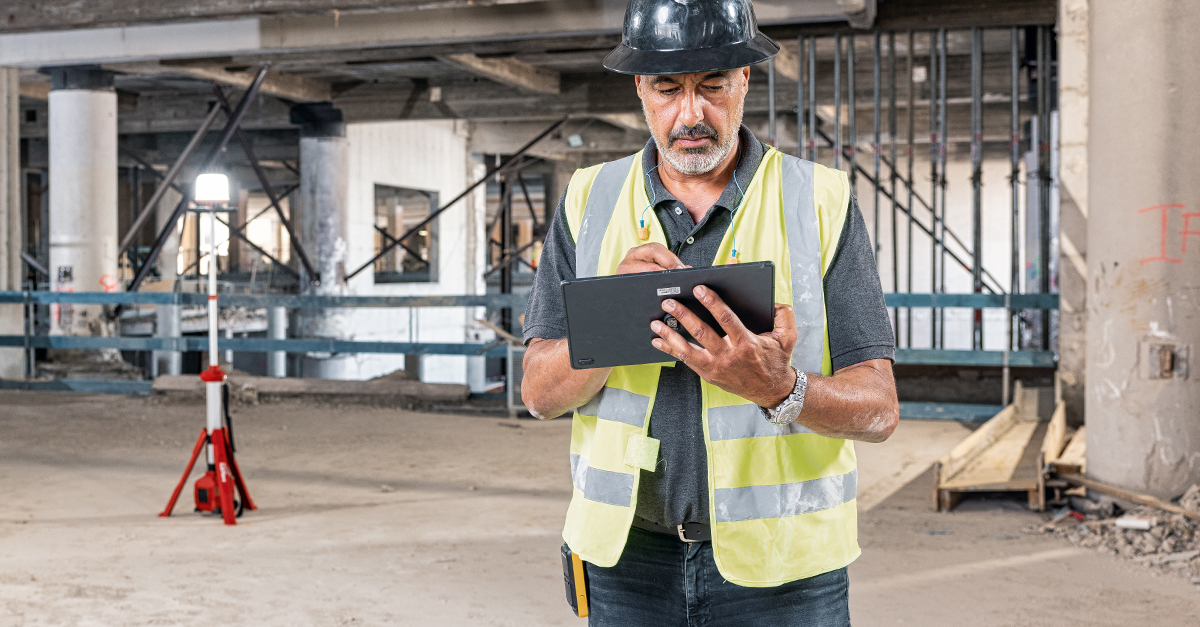 A construction workers in high-visibility jacket and hard hat creating a RFI on a tablet at an active building site, illustrating the use of Ai features in construction management software.