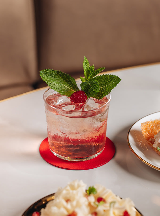 A glass with a pink drink, ice cubes, a sprig of mint, and a raspberry garnish on a red coaster on a table with food