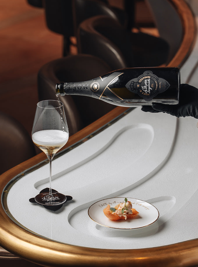 A bottle of Champagne being poured into a glass beside a small plated appetizer