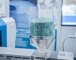 Ein transparenter Behälter mit hellblauen Kunststoffgranulaten steht auf einer metallischen Maschine in einer industriellen Umgebung.