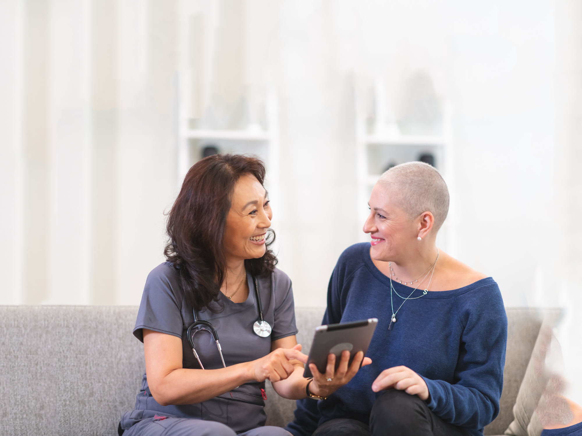 Hoitaja ja syöpää sairastava nainen istuvat vierekkäin sohvalla, hoitajalla tabletti käsissään. 