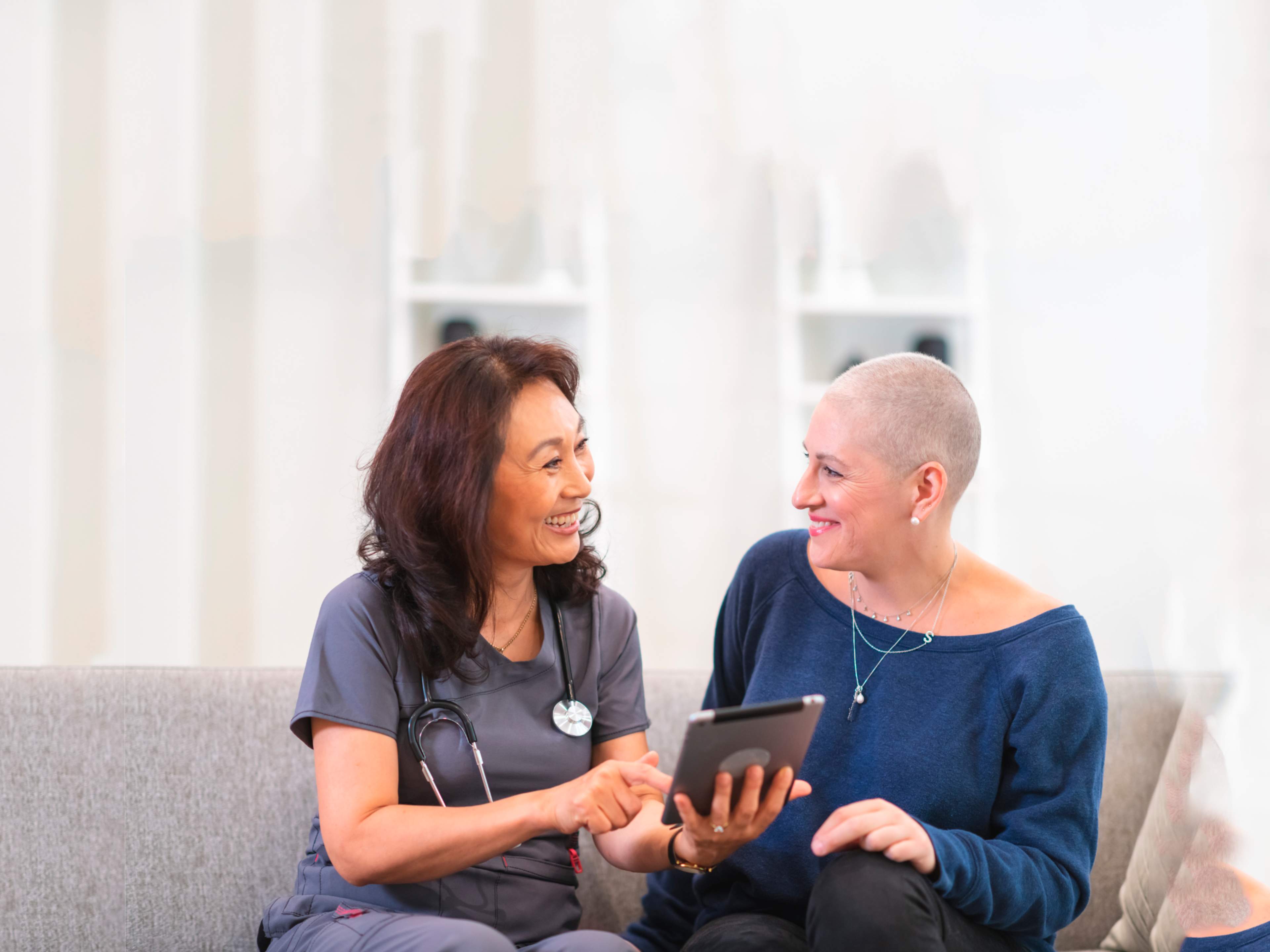 Hoitaja ja syöpää sairastava nainen istuvat vierekkäin sohvalla, hoitajalla tabletti käsissään.