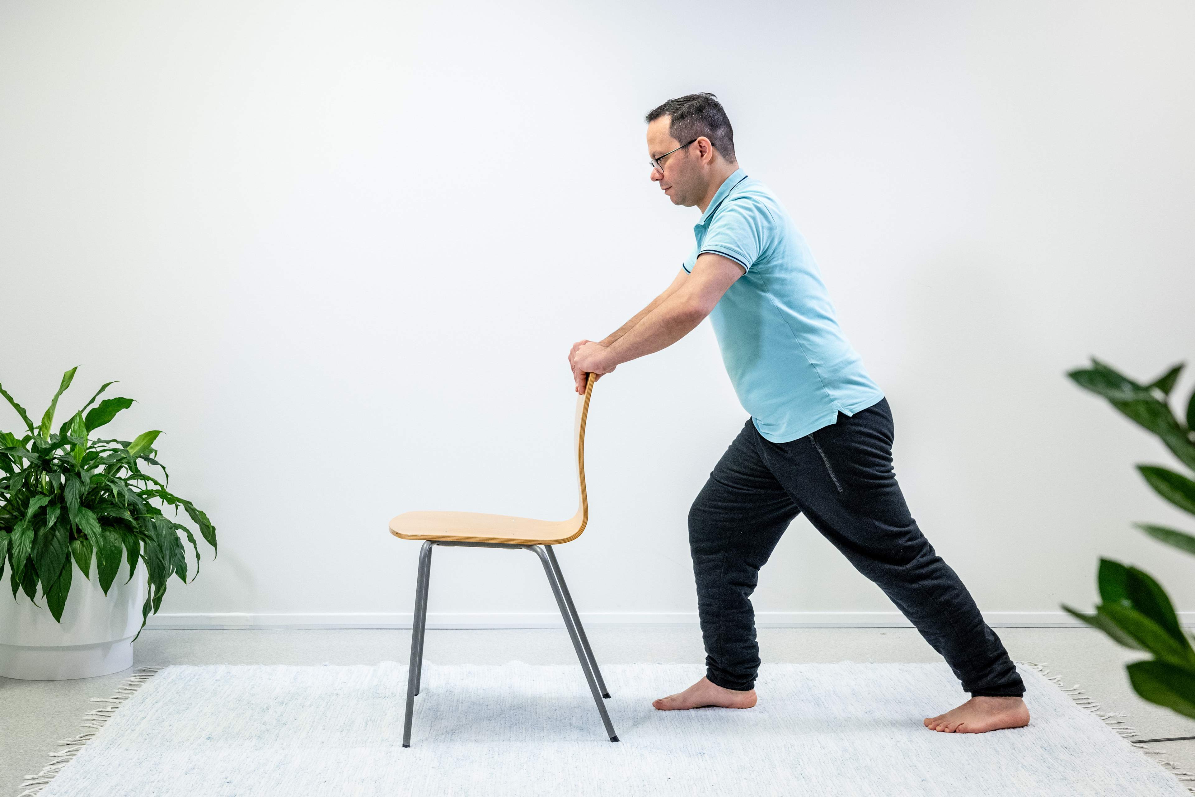 Mies pitää käsillä tukea tuolin selkänojasta ja on ottanut askeleen taaksepäin vasemmalla jalalla.