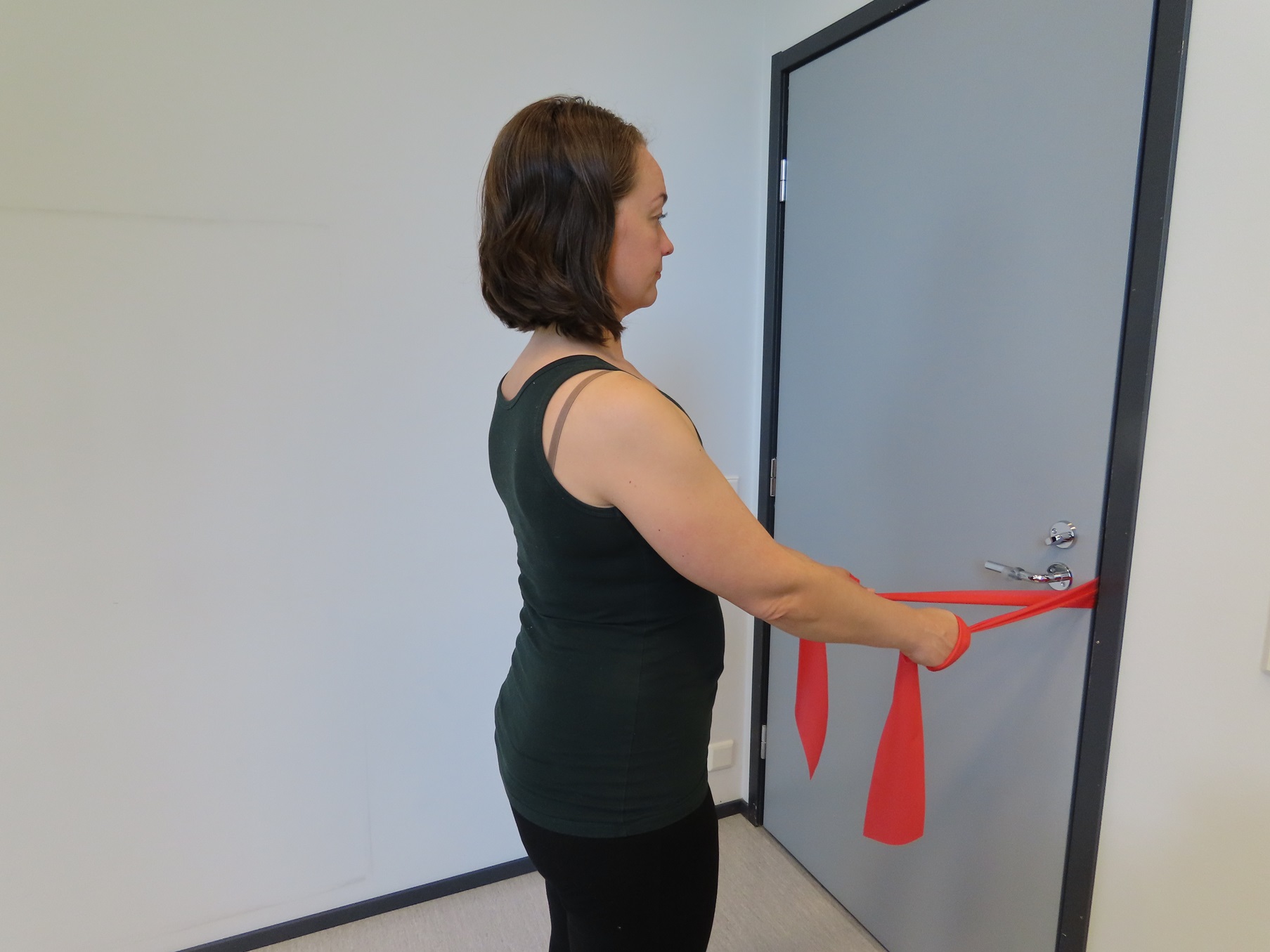 Nainen seisoo ovea kohti ja pitää vastusnauhasta kiinni molemmilla käsillä. Vastusnauha on kiinni oven kahvassa ja ovi on kiinni.