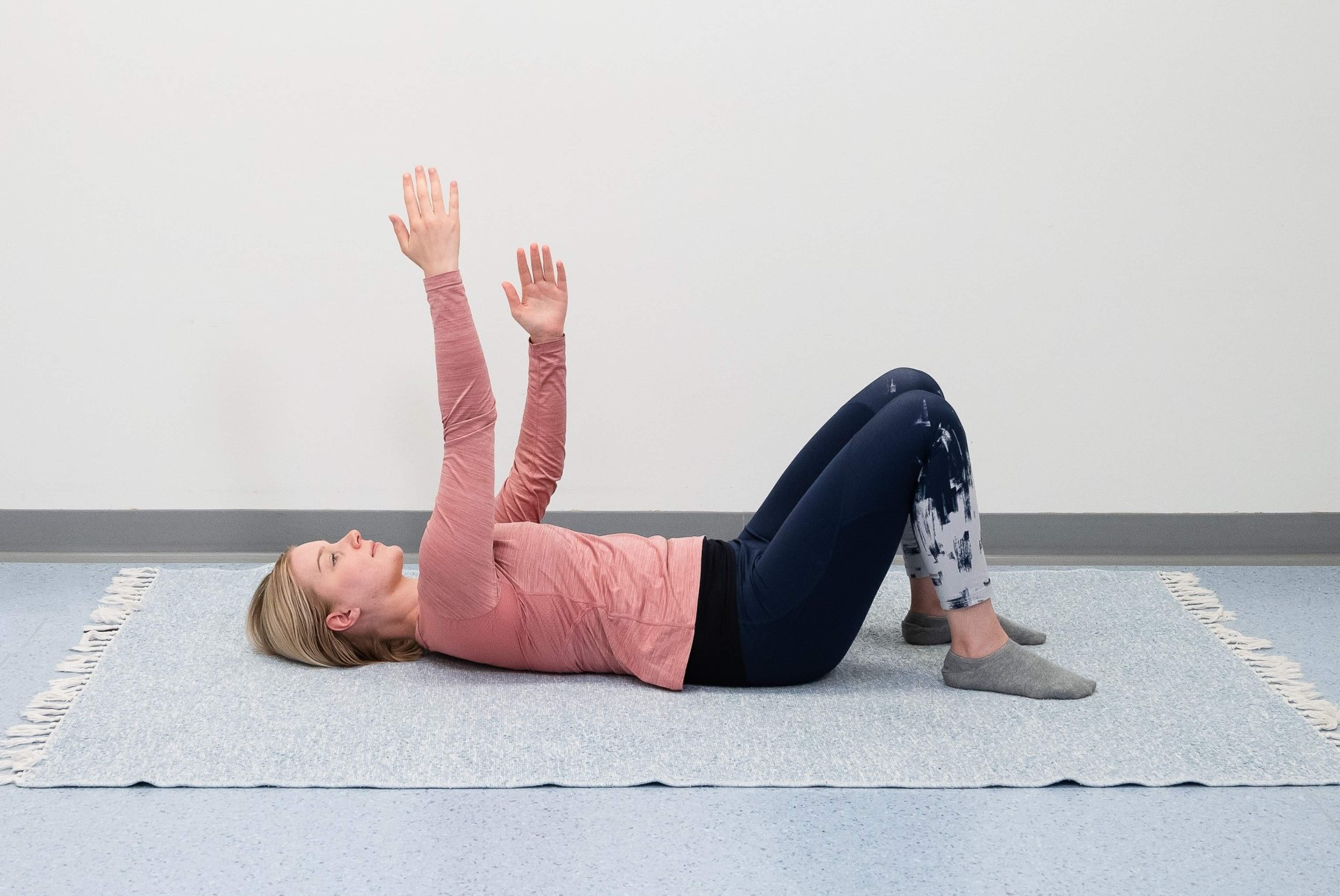 Nainen makaa selinmakuulla lattialla ja polvet koukussa. Nainen kurottaa toisella kädellä ylös kohti kattoa ja toinen käsi on suorana ylöspäin.