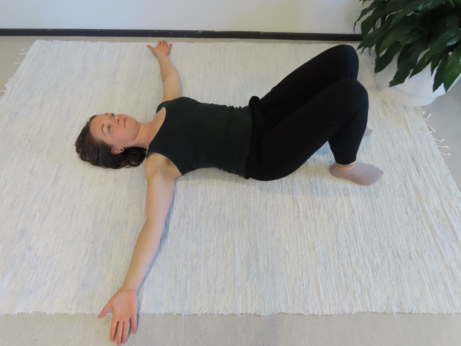 Nainen makaa selinmakuulla lattialla, polvet koukussa ja kädet vietynä lattiaa pitkin sivulle kämmenselät lattiaa vasten.