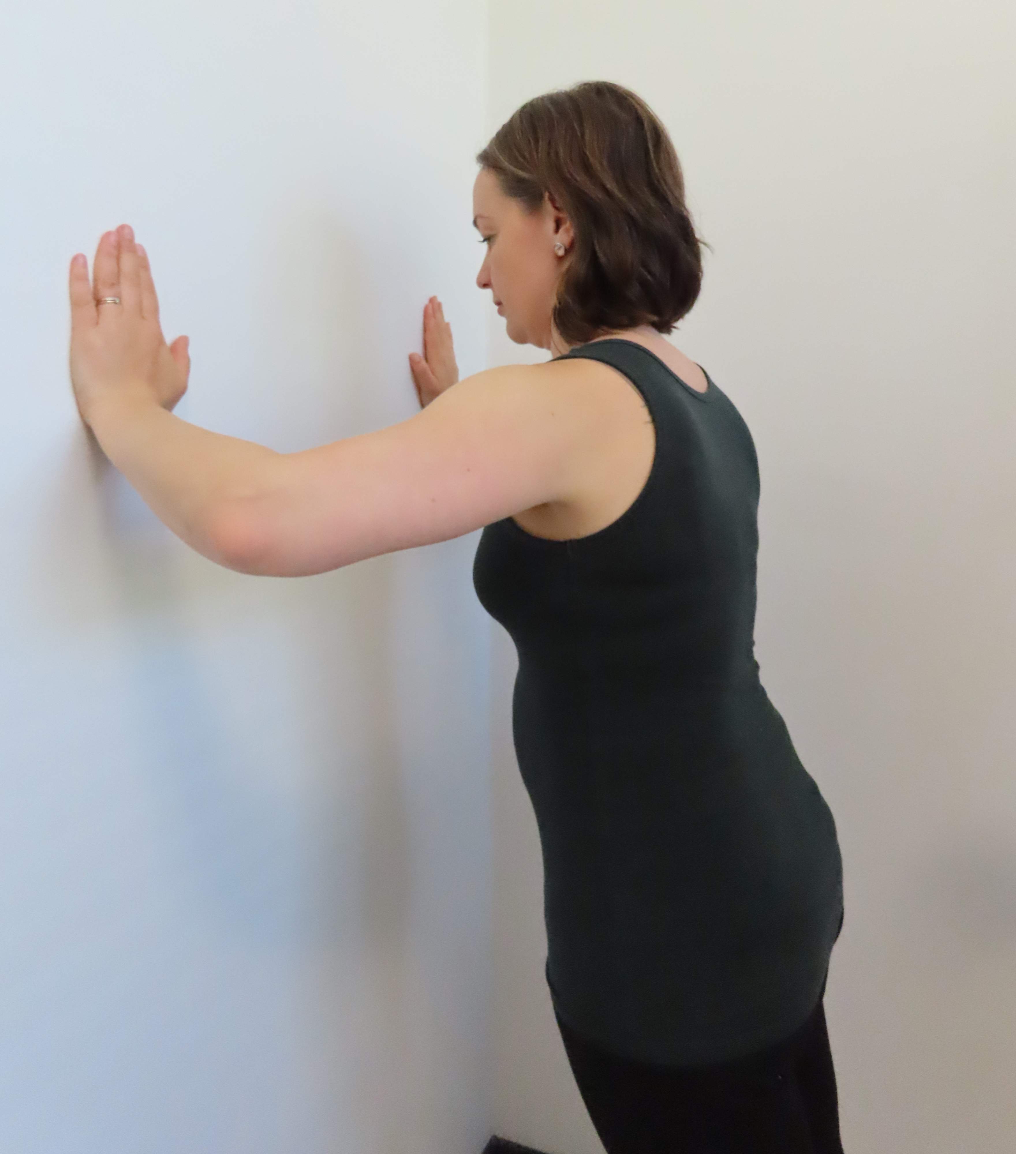 Nainen seisoo kasvot seinään päin, kädet leveässä punnerrusasennossa seinällä rinnan korkeudella, sormet osoittavat ylöspäin. Hän tekee rauhallisesti punnerrusliikettä vartalo suorana ja hartiat alhaalla.