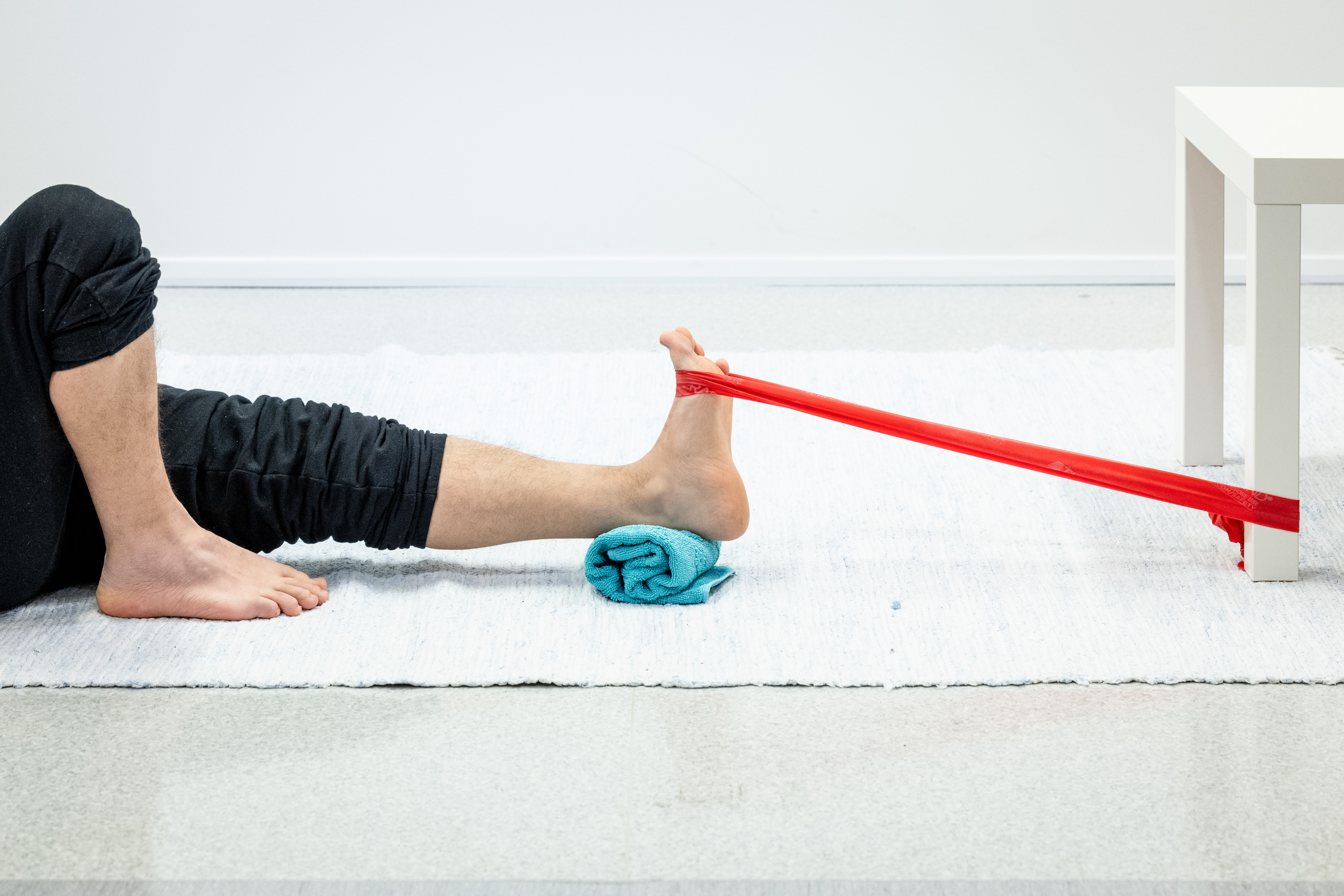 En man sitter på golvet med sitt vänstra ben rakt framför sig och en handduk rullad under fotleden. Motståndsbandet är runt mannens vänstra fotled och fäst vid ett bord. Mannen har böjt sin vänstra fotled.