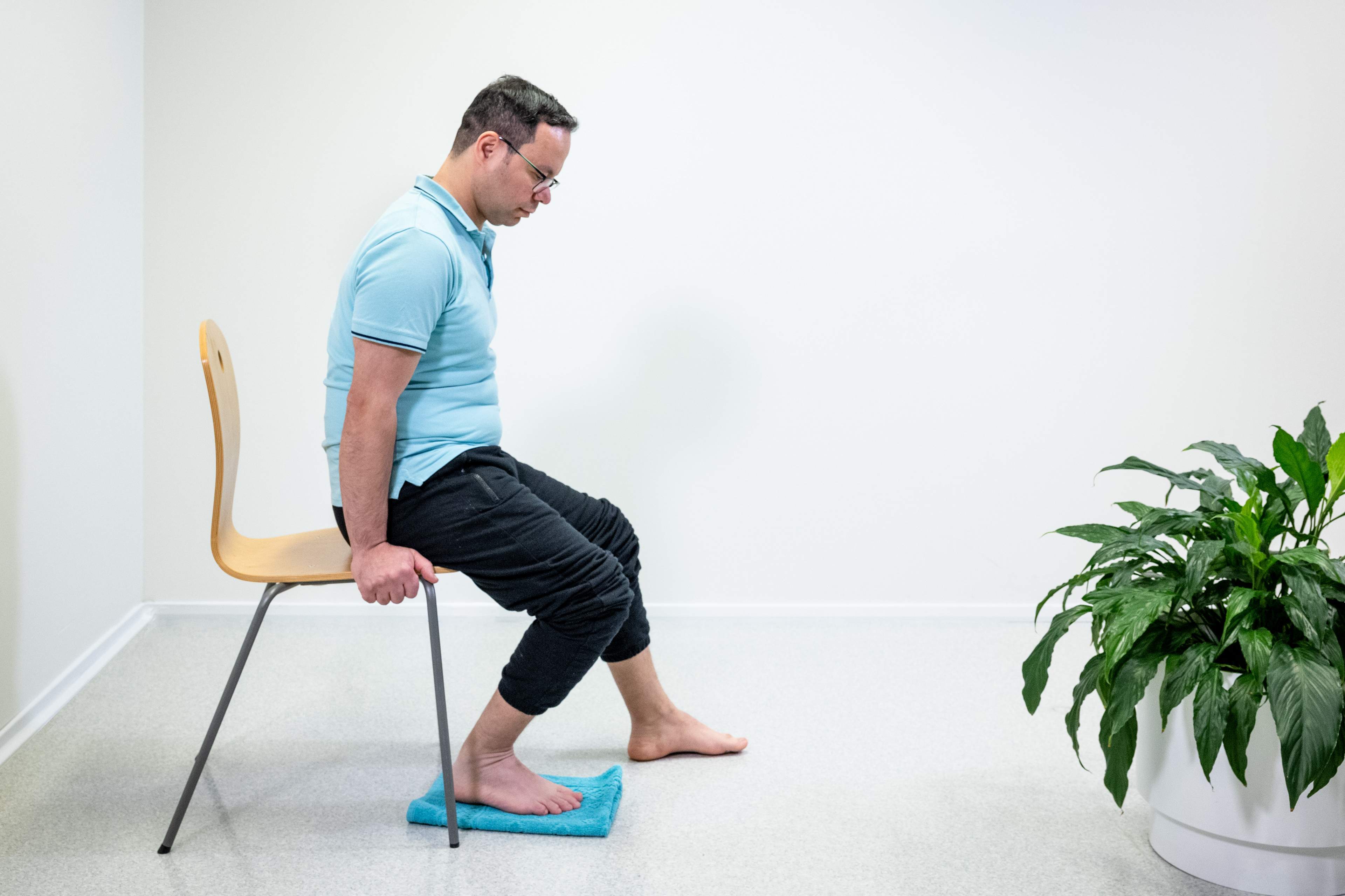 Mies istuu tuolin reunalla ja on vetänyt oikean jalan koukkuun tuolin alle. Kantapää on lattiassa ja jalan alla on pyyhe.