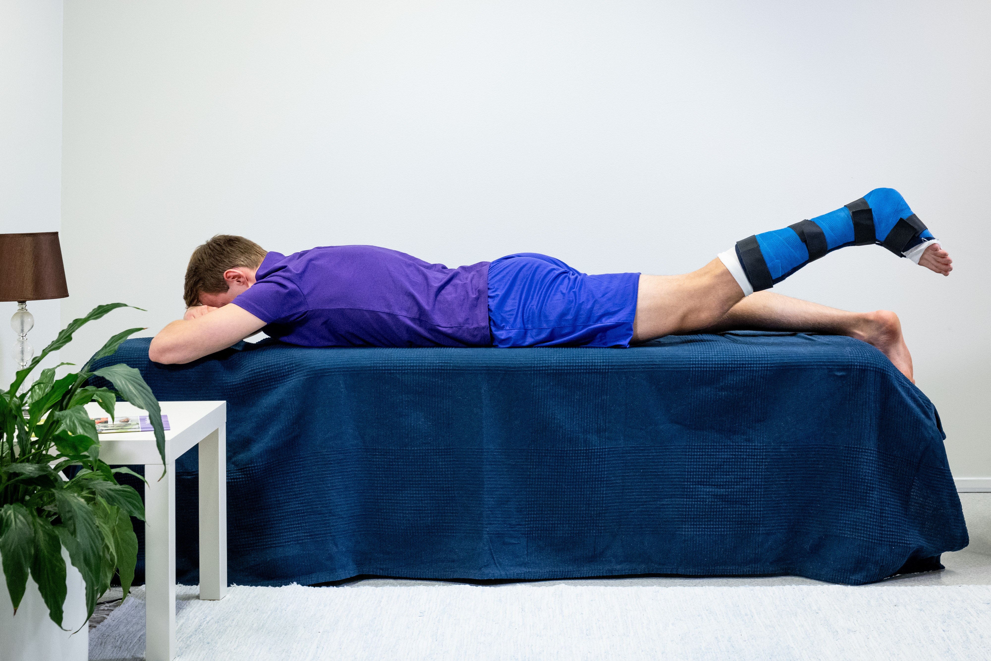 Mannen ligger på mage med vristerna utanför sängkanten, och det vänstra benet är i gips. Mannen har lyft det vänstra benet rakt uppåt med benet sträckt.