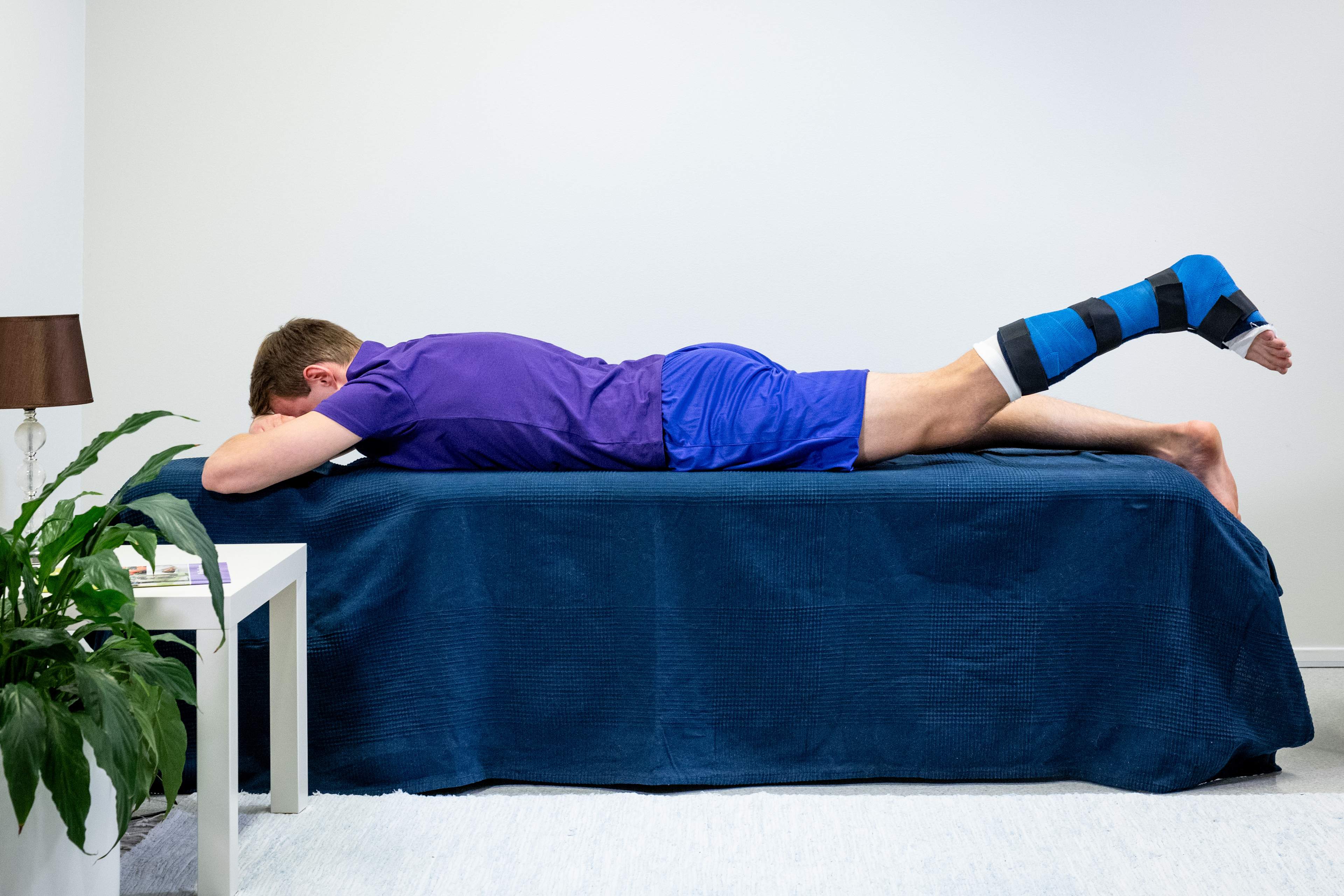 Mannen ligger på mage med vristerna utanför sängkanten, och det vänstra benet är i gips. Mannen har lyft det vänstra benet rakt uppåt med benet sträckt.