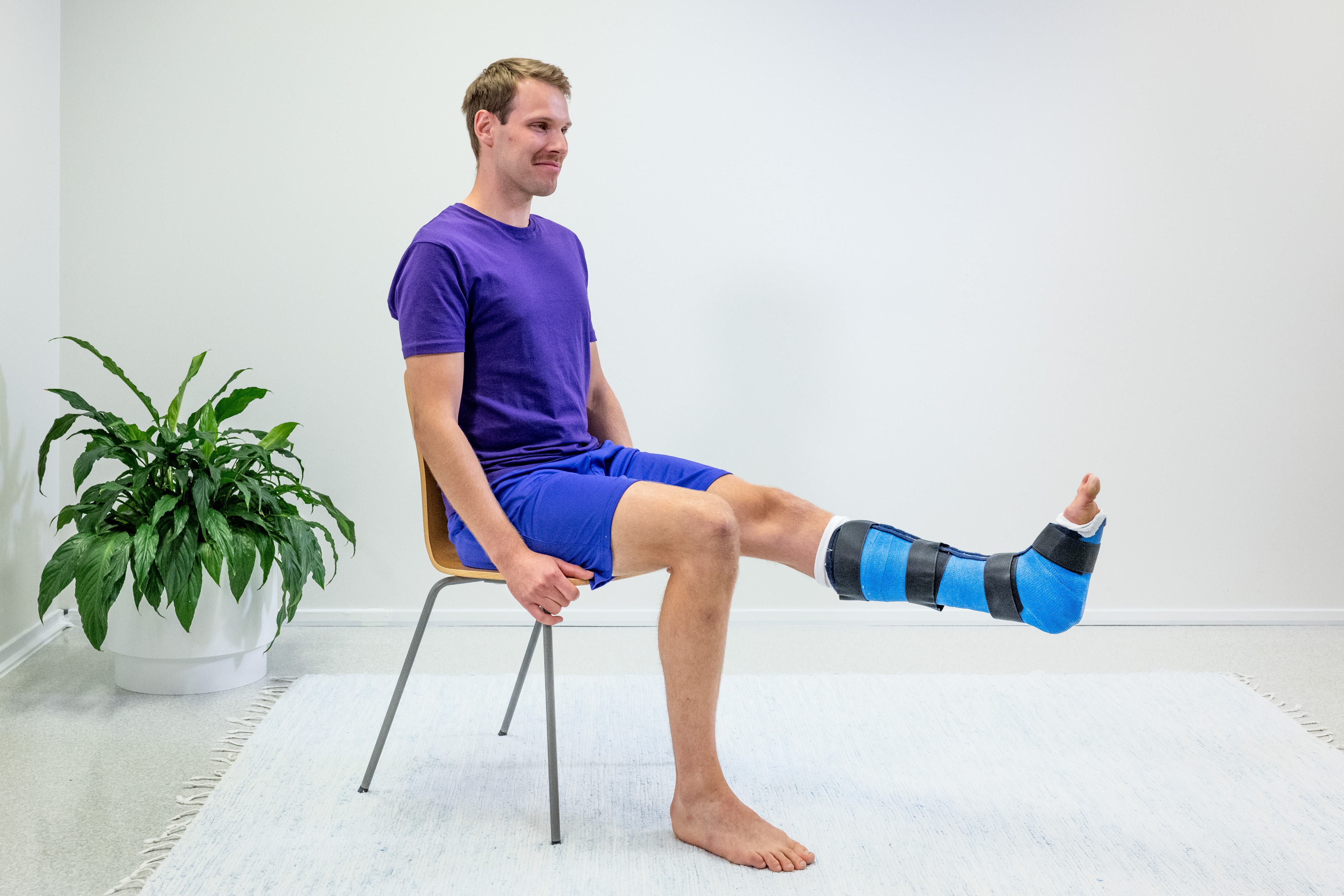 Mannen sitter på en stol med fötterna på golvet, och han har gips på vänster fotled. Mannen har sträckt ut knäet på det gipsade benet.