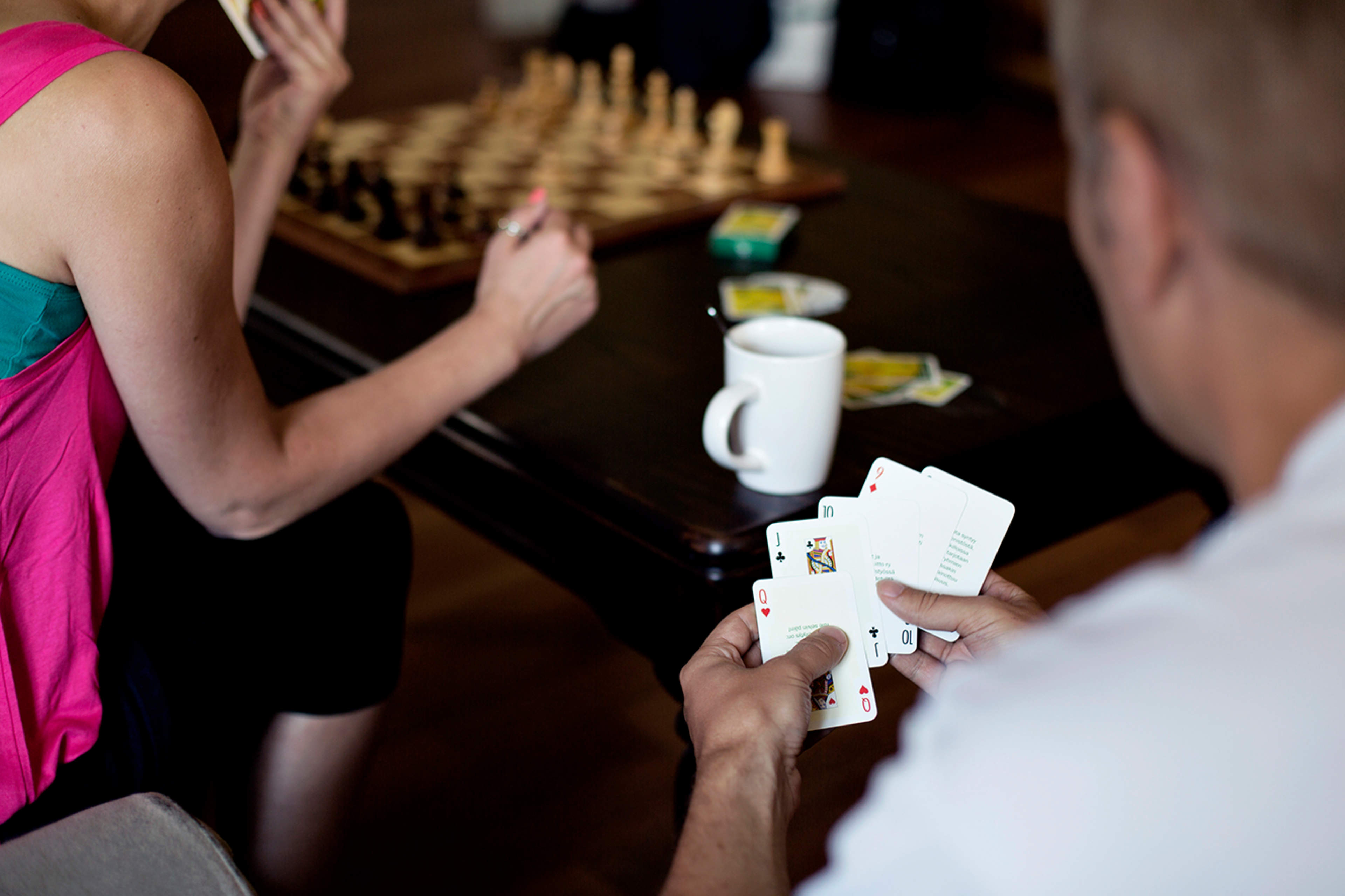 Shakkia ja korttipeliä tumman pöydän äärellä. Kaksi ihmistä pöydän äärellä selin kuvaajaan ja vain osittain kuvassa