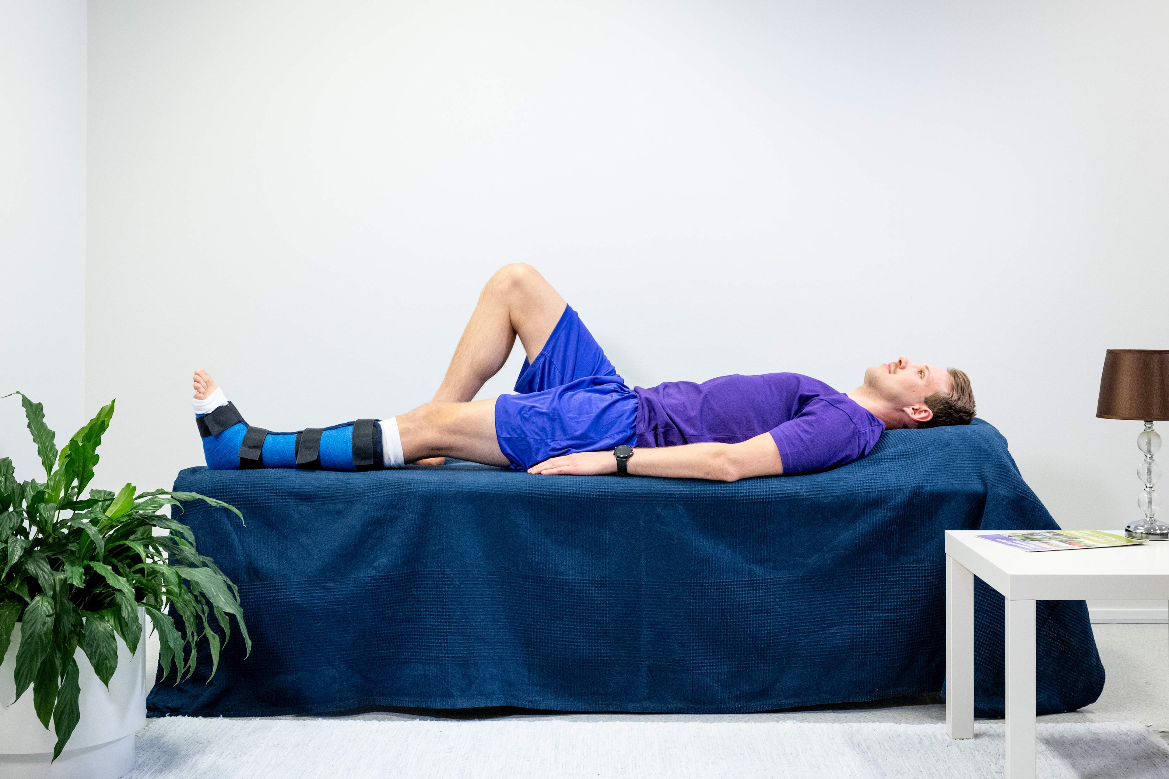 Mannen ligger på rygg i sängen med ett gips på vänster fotled, och det högra benet är böjt på sängen.