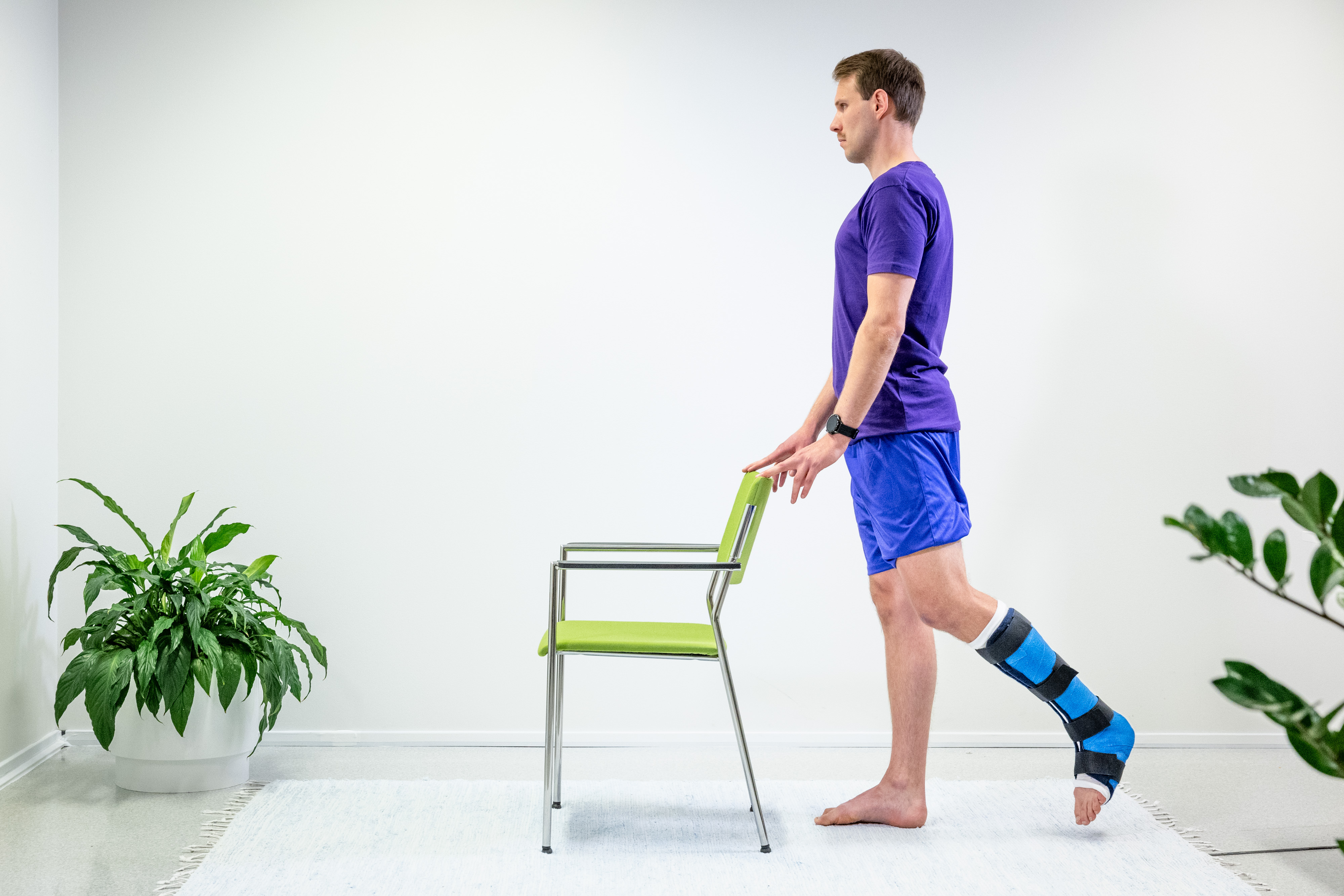 Mies seisoo ja pitää kiinni edessä olevan tuolin selkänojasta, vasen nilkka on kipsattu. Mies on nostanut vasenta jalkaa taakse. 