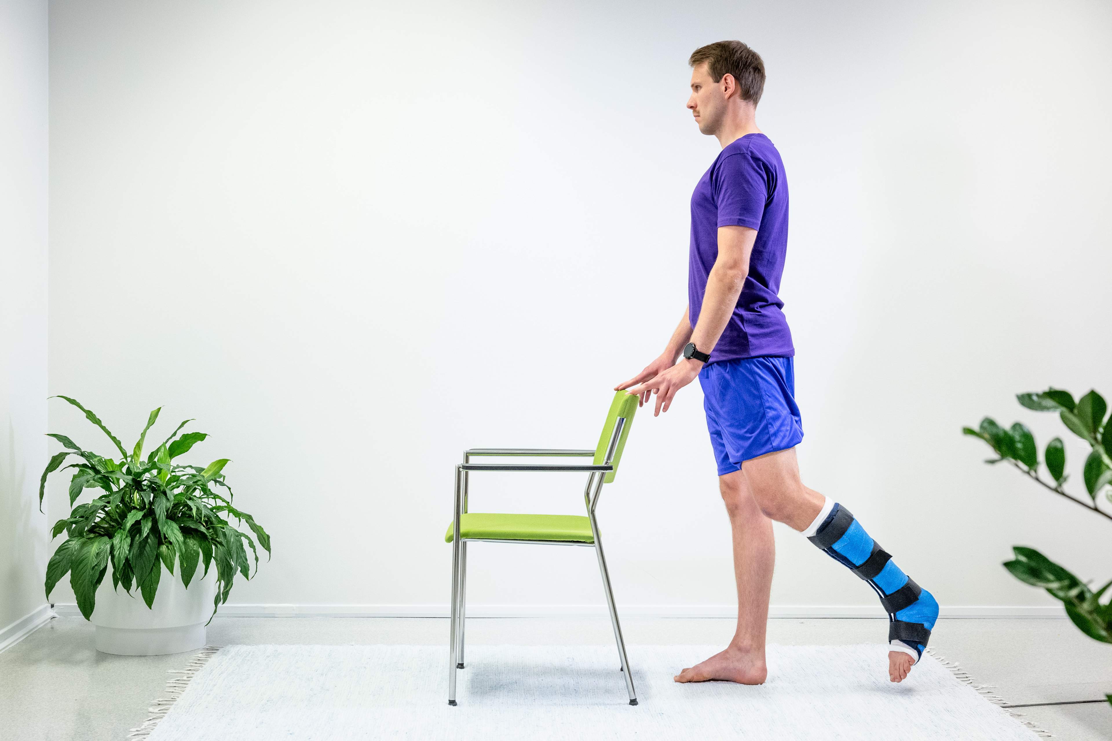 Mannen står och håller i ryggstödet på en stol framför sig, och det vänstra benet är gipsat. Mannen har lyft det vänstra benet bakåt.
