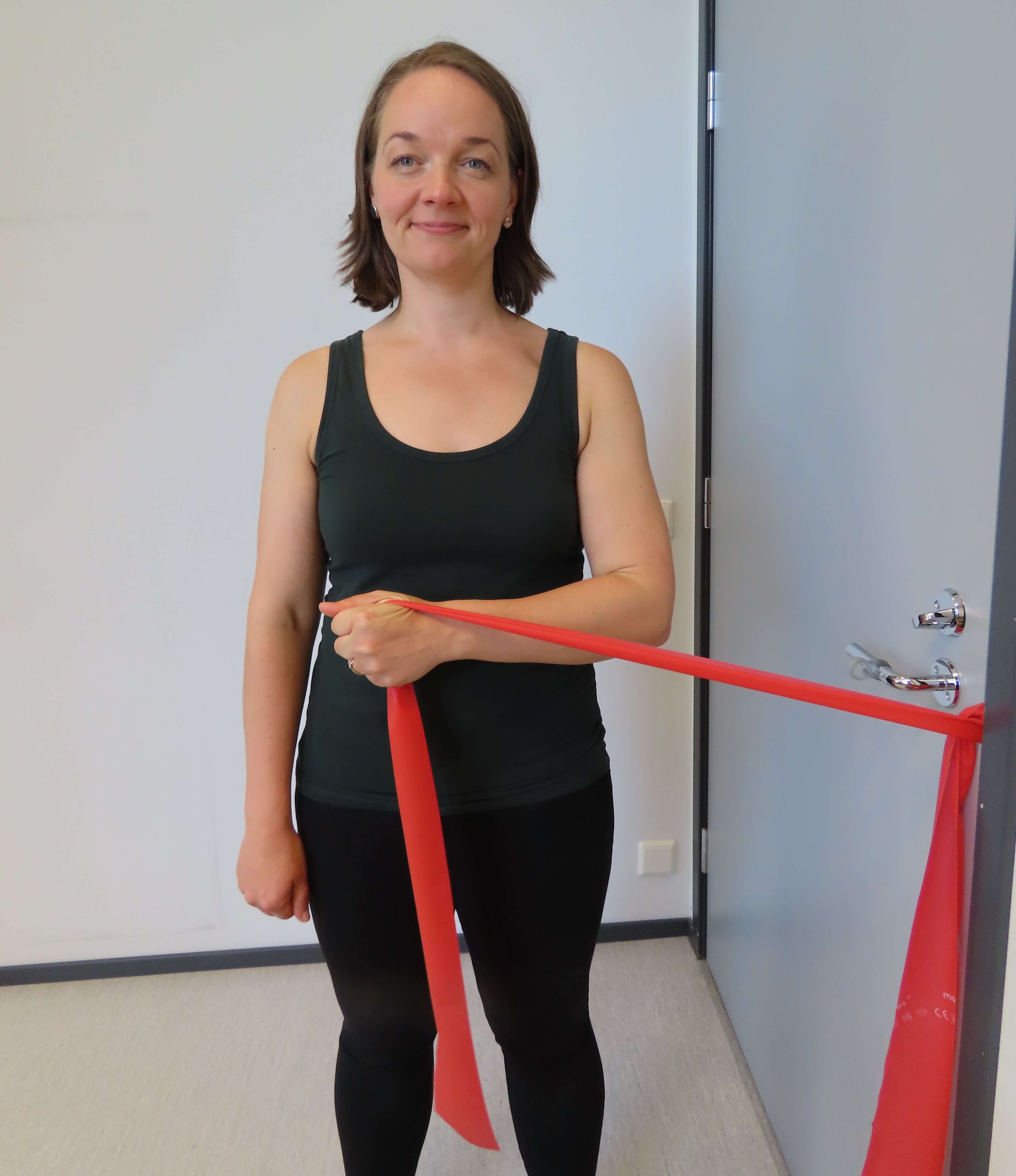 Kvinnan för handen inåt med armbågen böjd och intill sidan. Kvinnan håller i ett motståndsband. Motståndsbandet är fäst i dörrhandtaget och dörren är stängd.