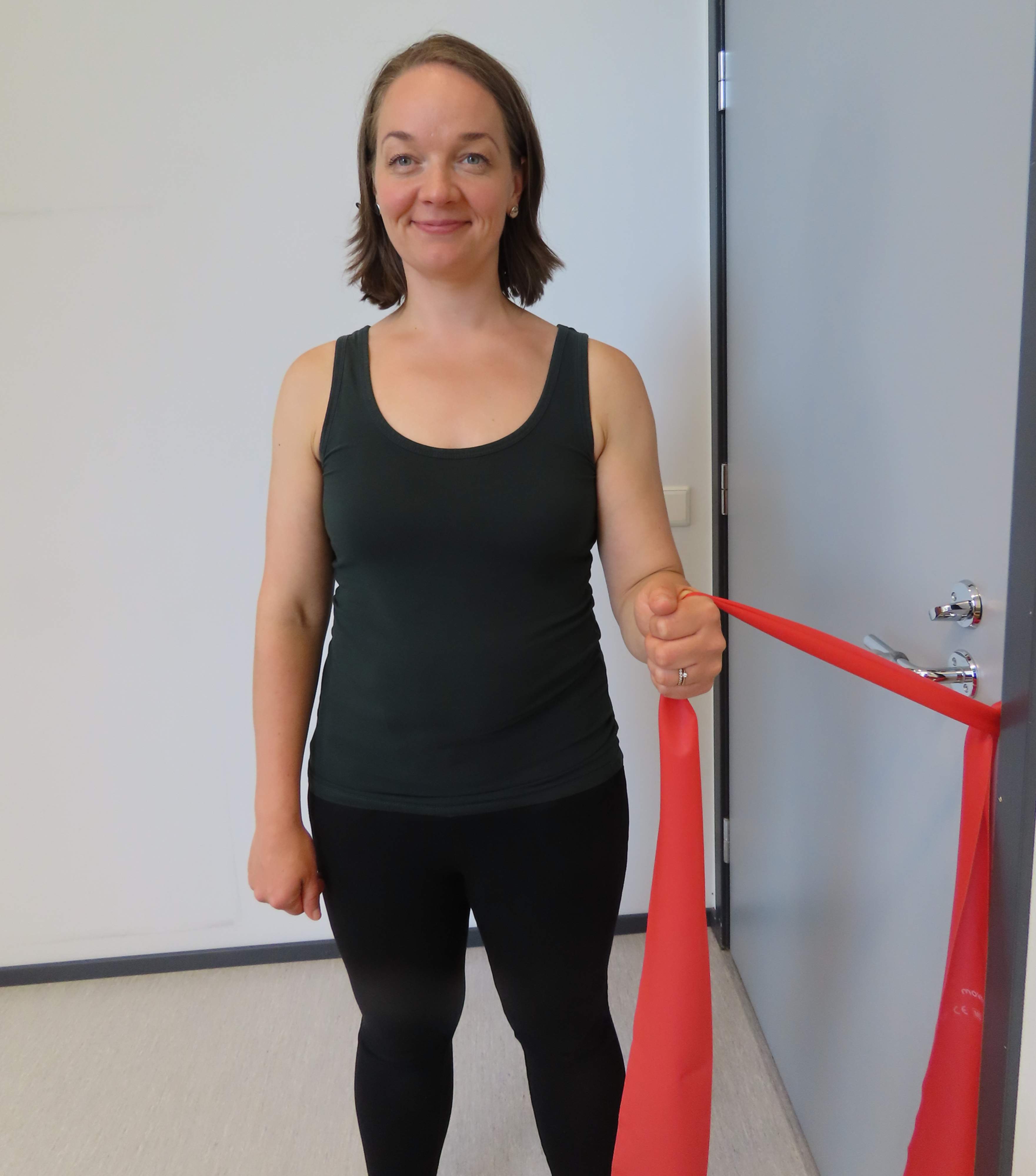 Nainen seisoo kyynärpää koukussa kylki ovea kohti ja pitää vastusnauhasta kiinni. Vastusnauha on kiinni oven kahvassa ja ovi on kiinni.