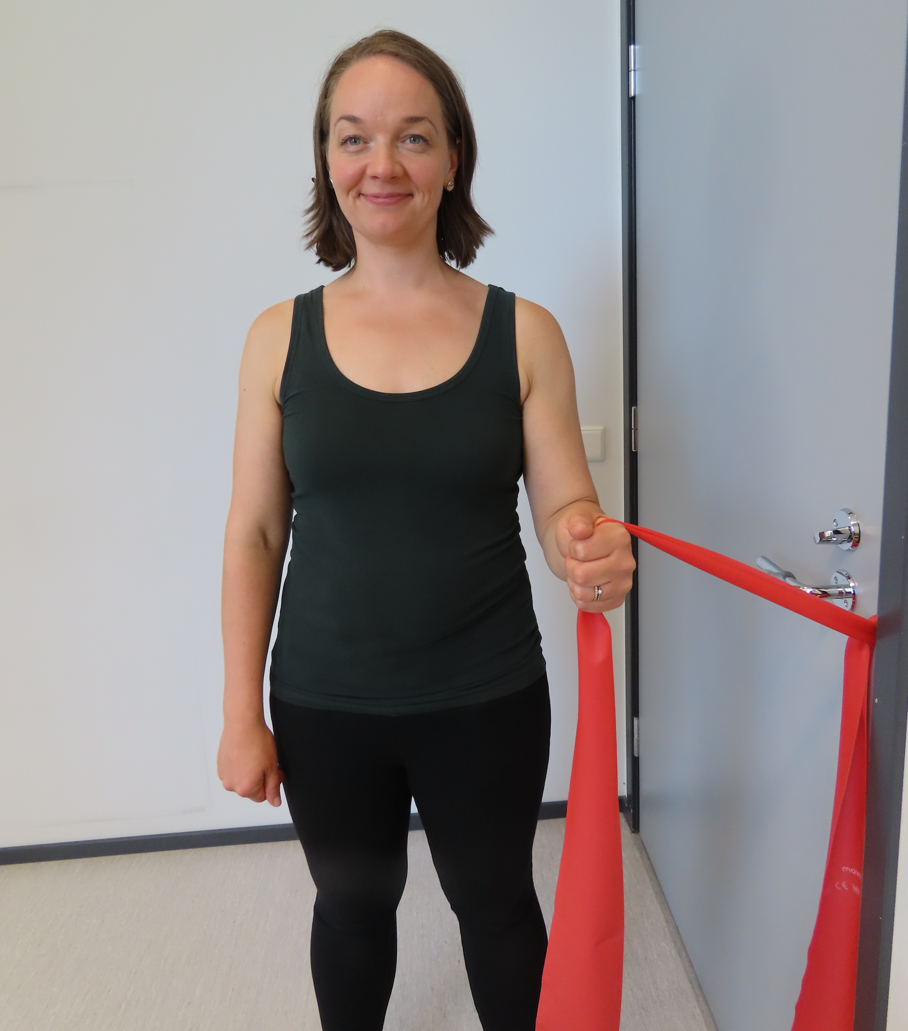 Kvinnan står med armbågen böjd och sidan av kroppen vänd mot dörren och håller i ett motståndsband. Motståndsbandet är fäst i dörrhandtaget och dörren är stängd.