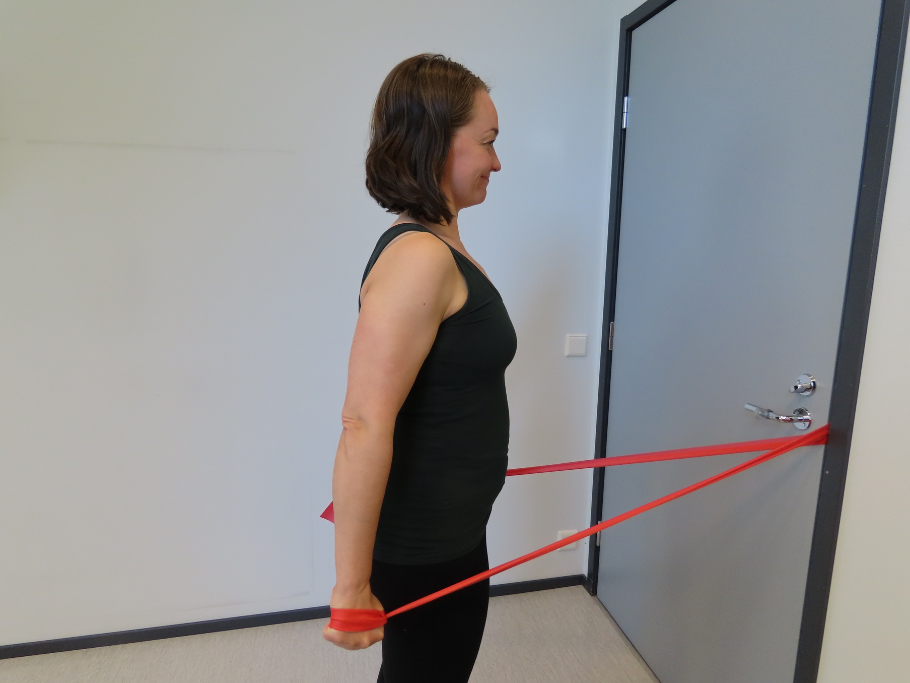 Nainen seisoo ovea kohti ja vetää kädet vartalon vierelle. Nainen pitää vastusnauhasta kiinni molemmilla käsillä. Vastusnauha on kiinni oven kahvassa ja ovi on kiinni.