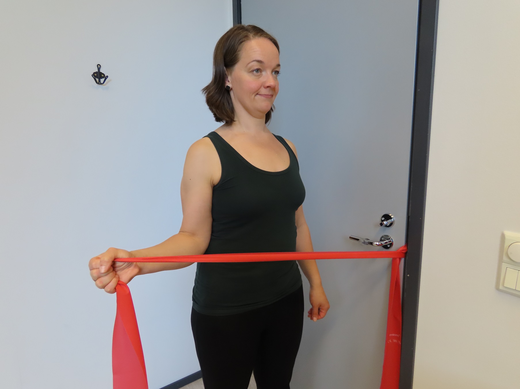 Kvinnan för underarmen utåt med armbågen böjd och tätt intill sidan. Kvinnan håller i motståndsbandet. Motståndsbandet är fäst i dörrhandtaget och dörren är stängd.