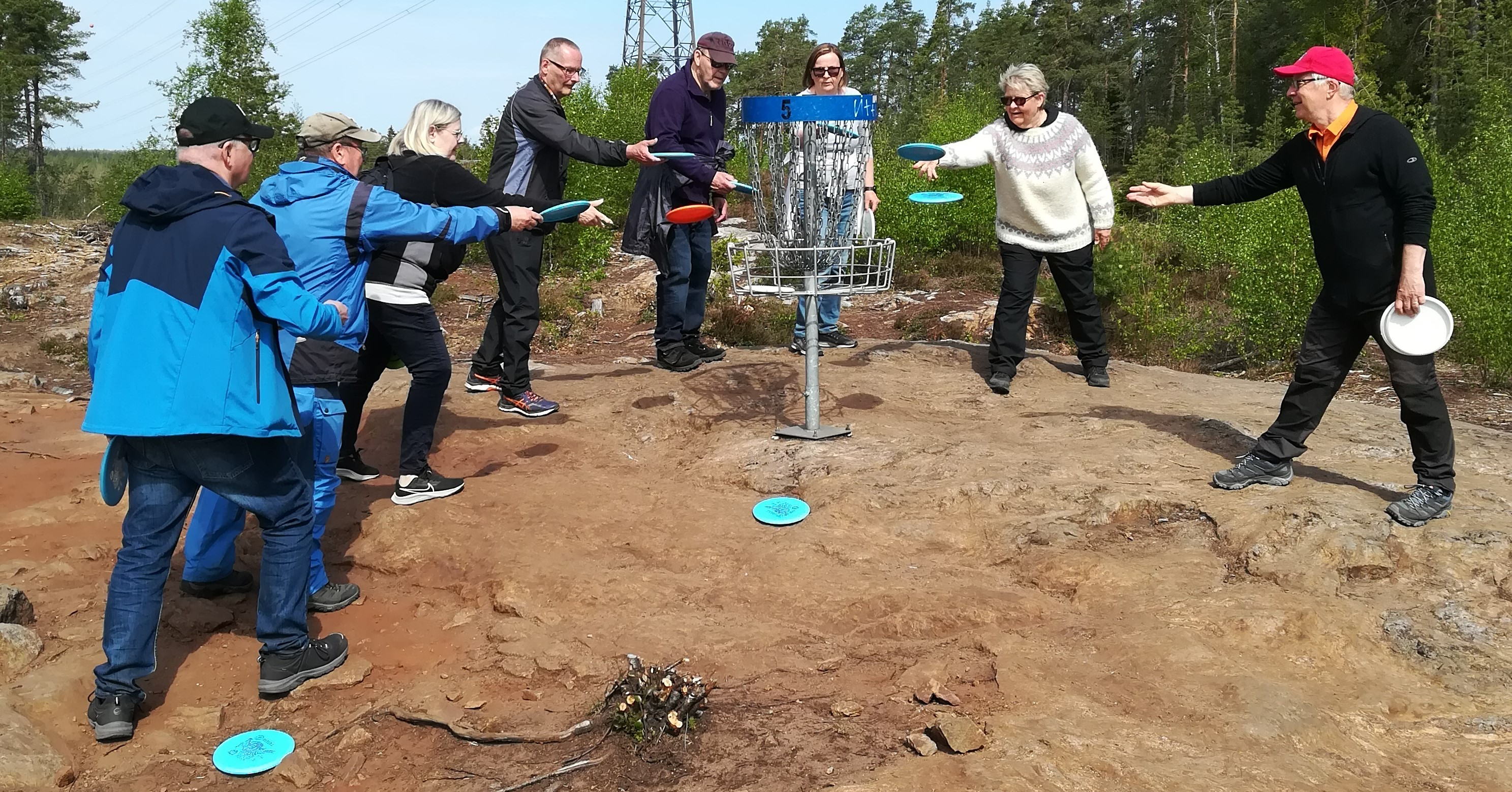 Kahdeksan henkilöä heittää frisbeegolf-kiekkoja samaan koriin kallioisessa maisemassa.