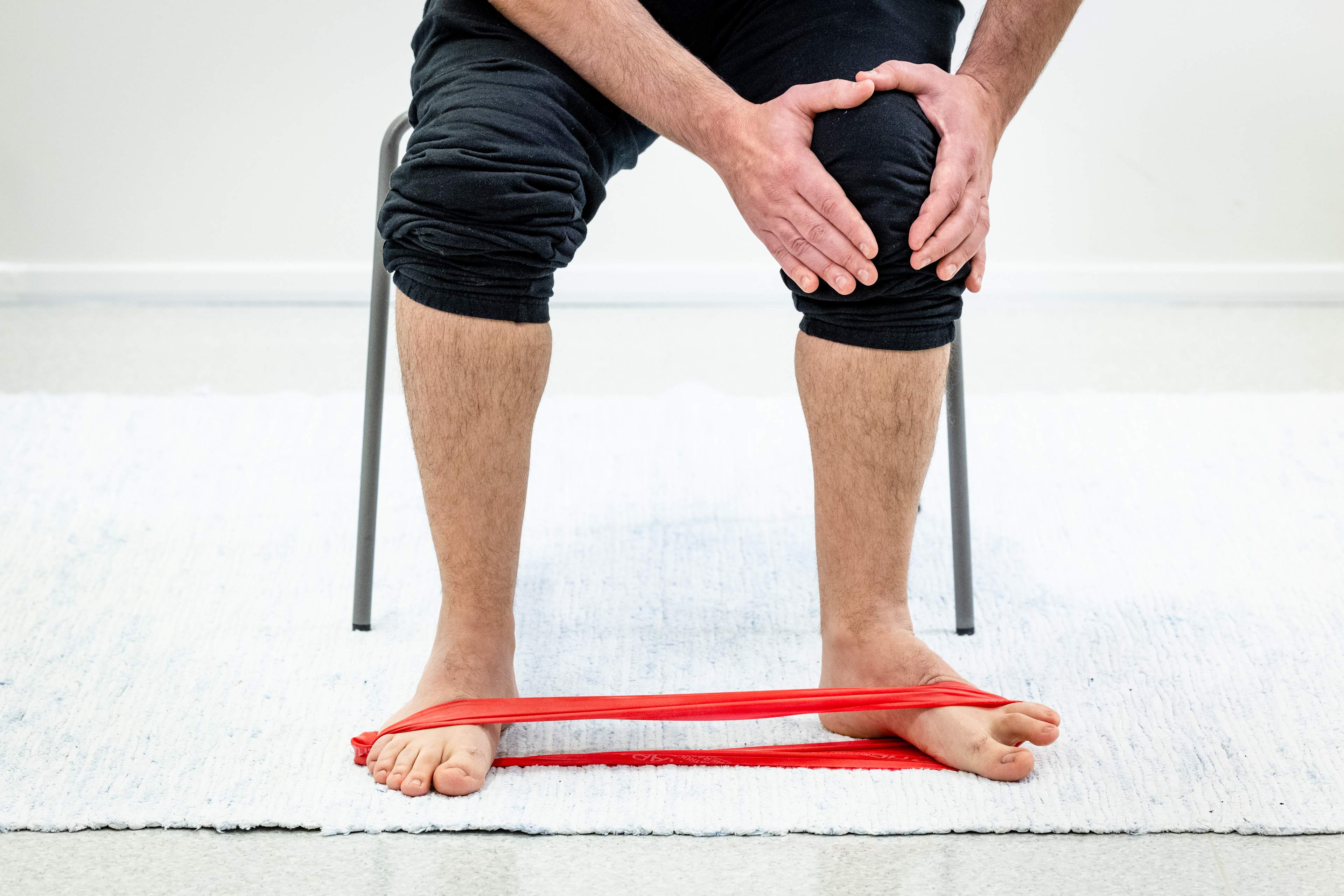 En man sitter på en stol och håller i sina händer vid vänster knä, med motståndsbandet runt fötterna. Mannen har vridit sin vänstra fot utåt mot lilltån, medan hälen har stannat på plats.