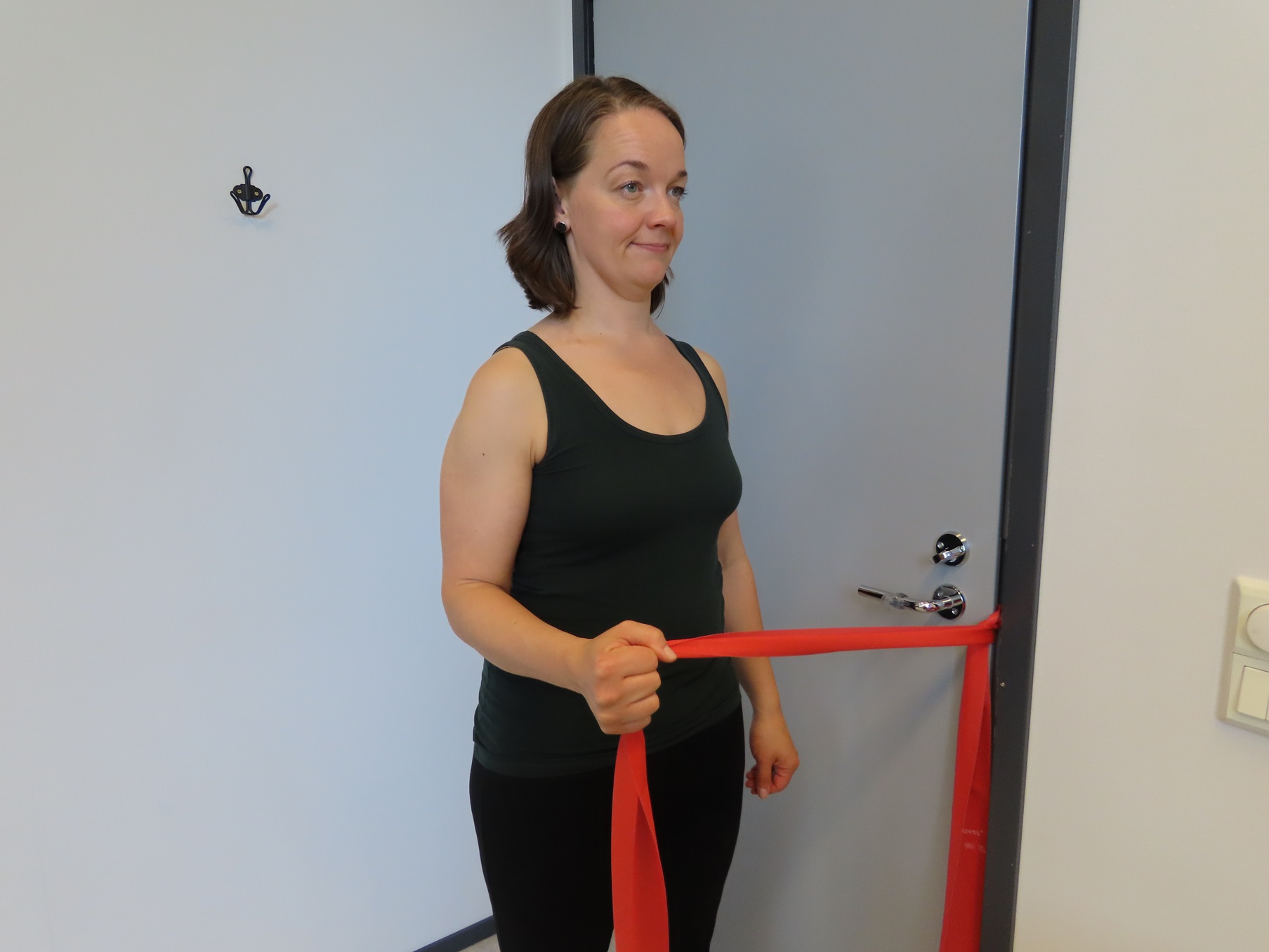 Kvinnan står med armbågen böjd, vänster sida av kroppen vänd mot dörren och håller i motståndsbandet med höger hand. Motståndsbandet är fäst i dörrhandtaget och dörren är stängd.