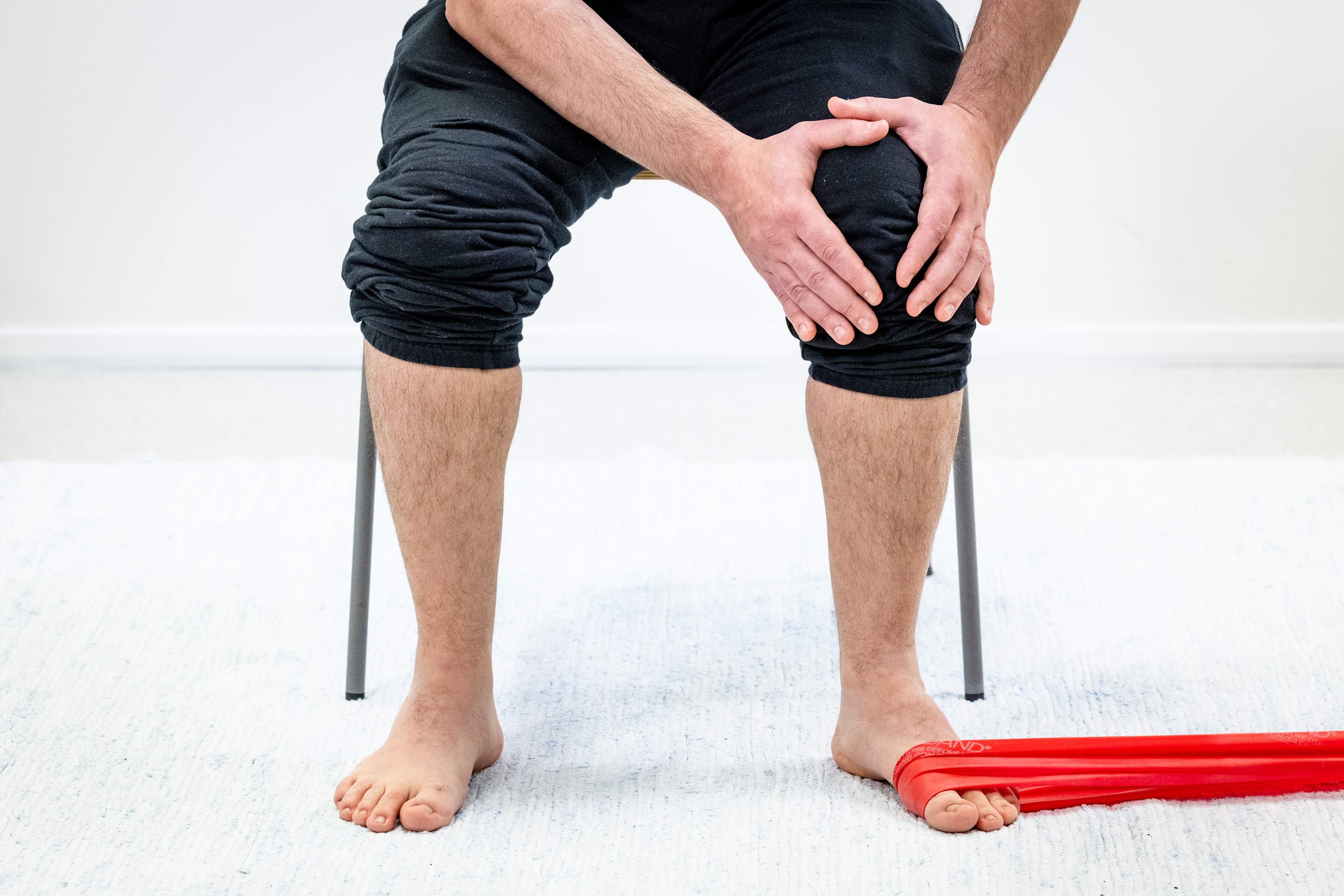 En man sitter på en stol och håller i sina händer vid vänster knä, med motståndsbandet runt vänster fot och fäst vid till exempel ett bord.