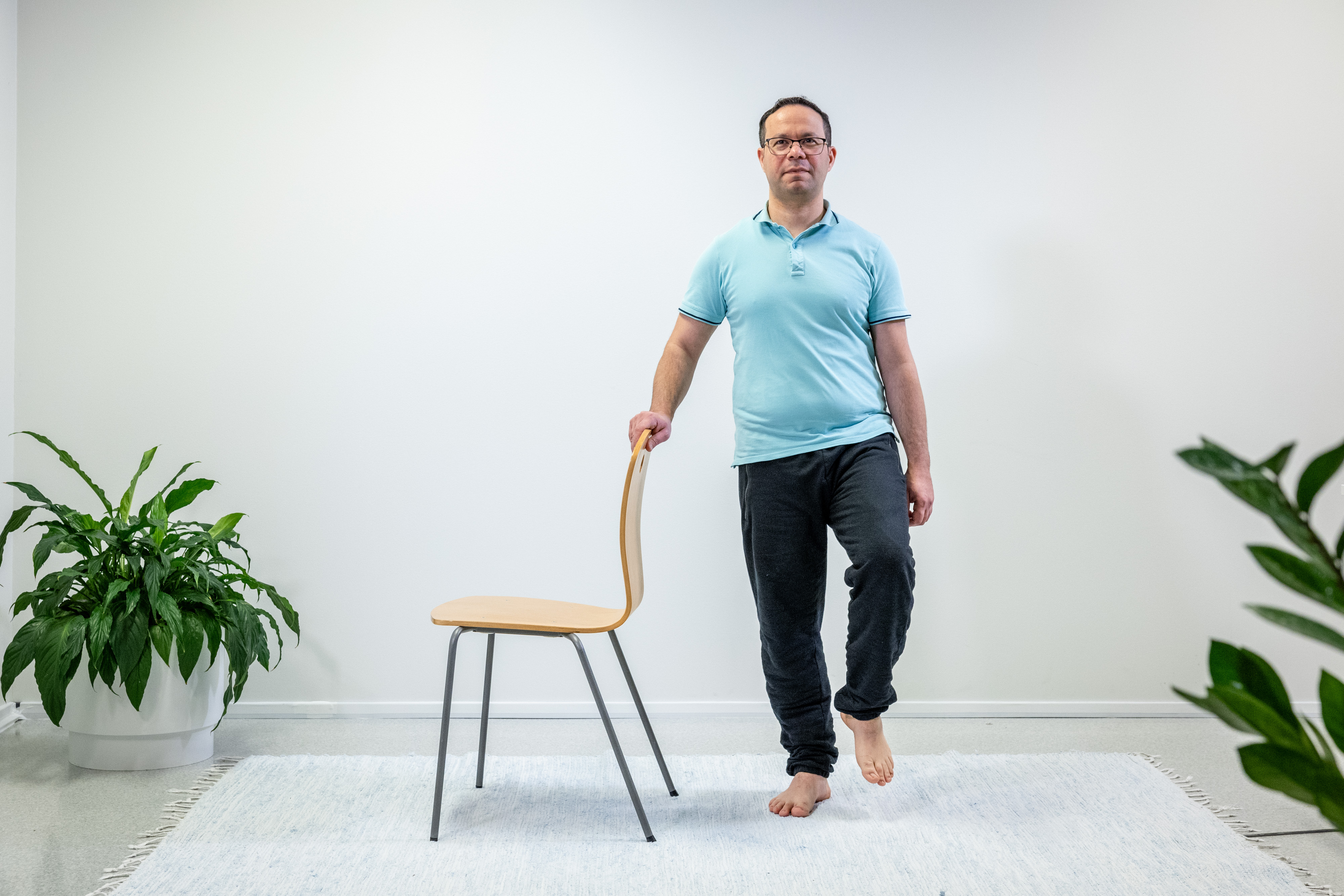 Mies pitää oikealla kädellä tukea tuolin selkänojasta ja seisoo yhdellä jalalla. Mies on nostanut vasemman jalan ilmaan. 