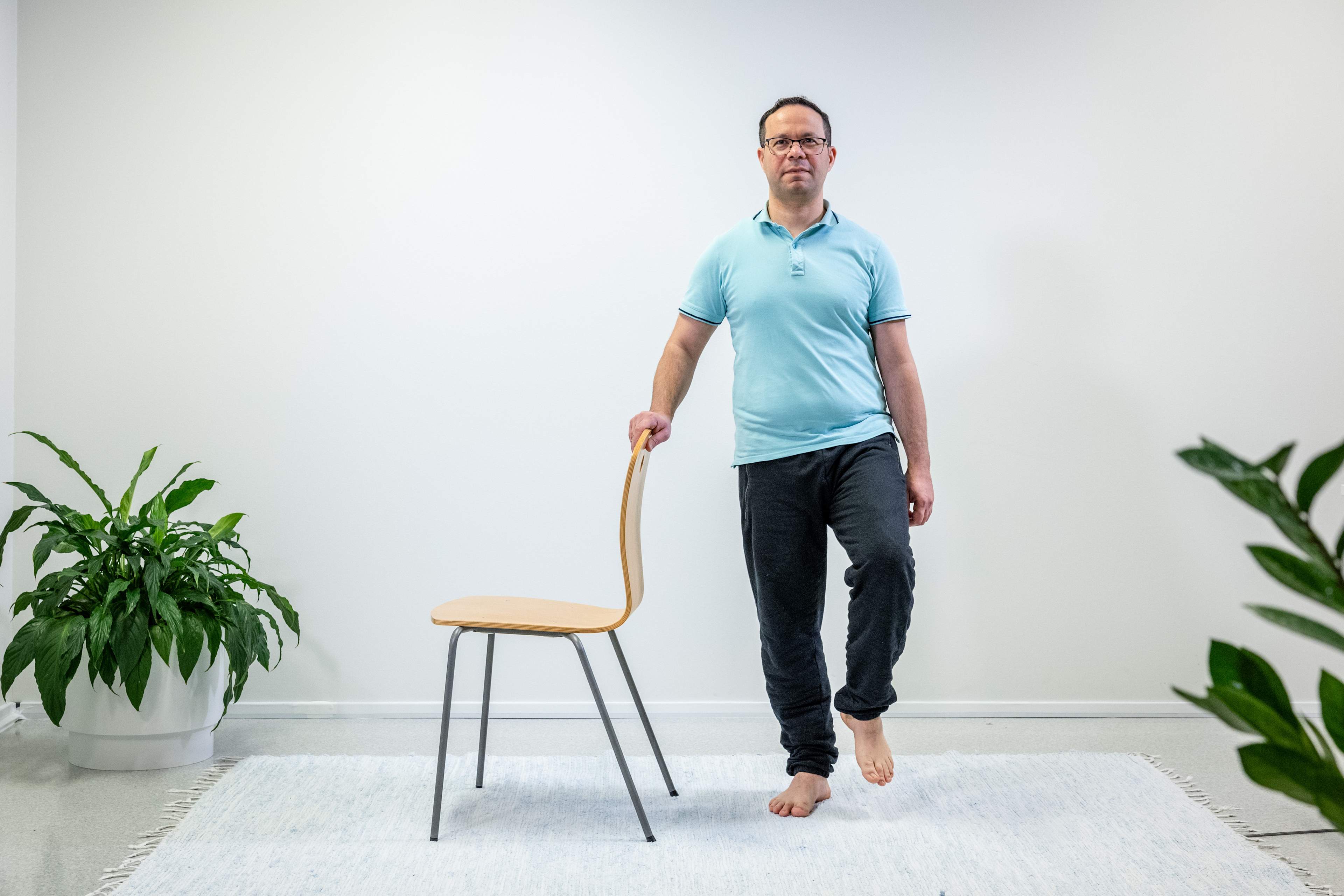 Mies pitää oikealla kädellä tukea tuolin selkänojasta ja seisoo yhdellä jalalla. Mies on nostanut vasemman jalan ilmaan.