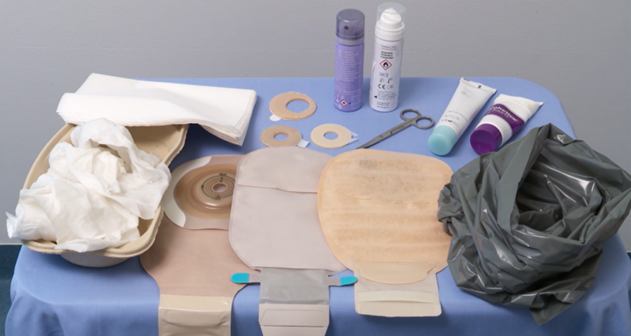 På bordet finns stomiförbandsartiklar: endelsbandage, limborttagningssrayer, pastaringar, tätningspasta, sax, soppåse, fuktiga och torra pappershanddukar.