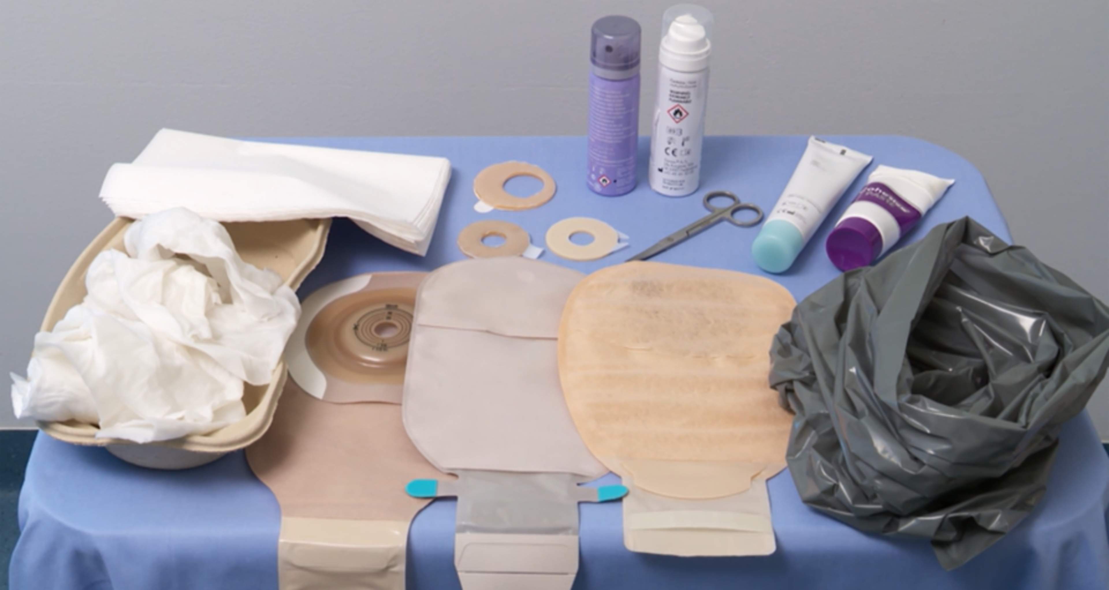 På bordet finns stomiförbandsartiklar: endelsbandage, limborttagningssrayer, pastaringar, tätningspasta, sax, soppåse, fuktiga och torra pappershanddukar.