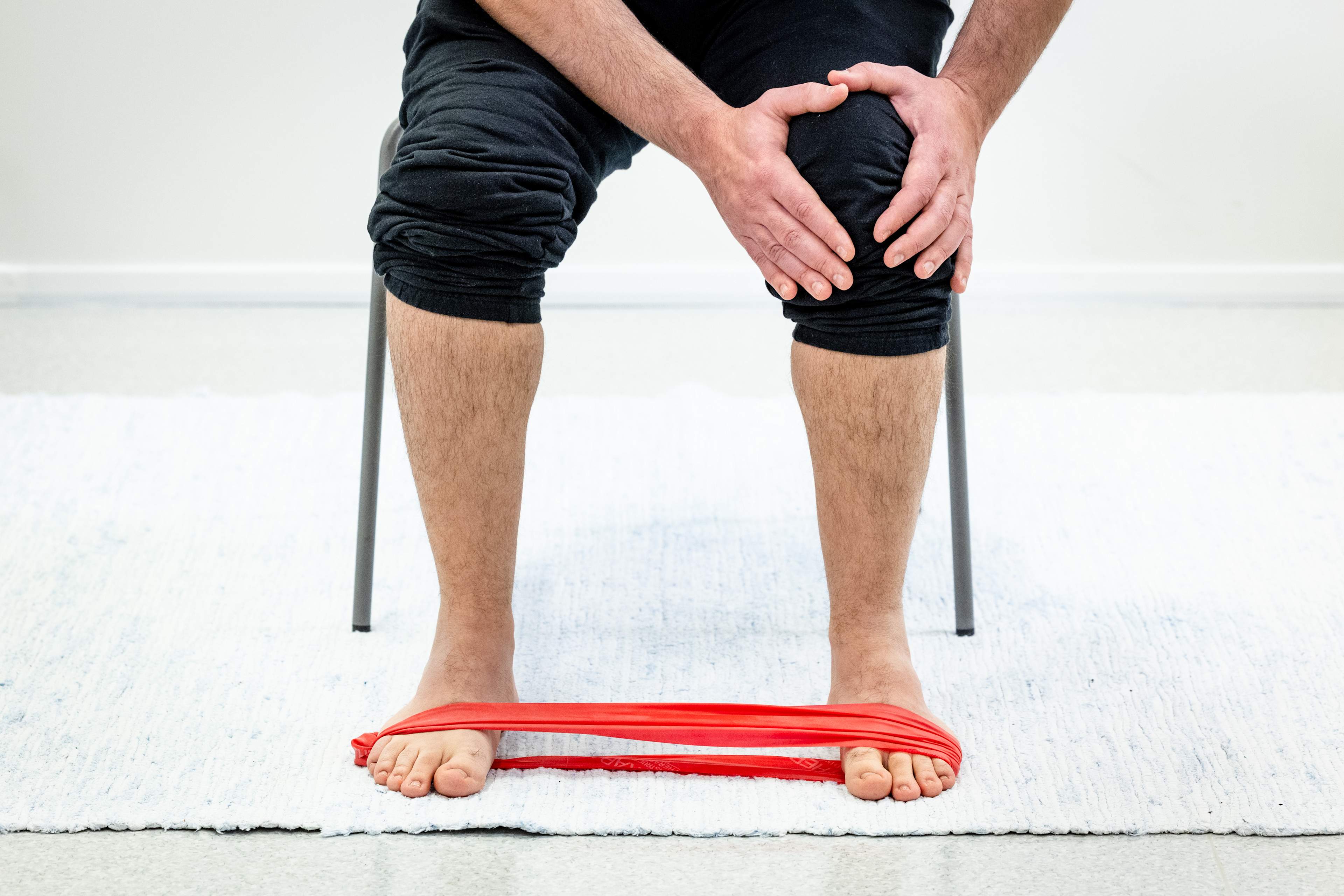 Mies istuu tuolilla ja pitää käsillä vasemmasta polvesta kiinni, vastuskuminauha on jalkaterien ympärillä.