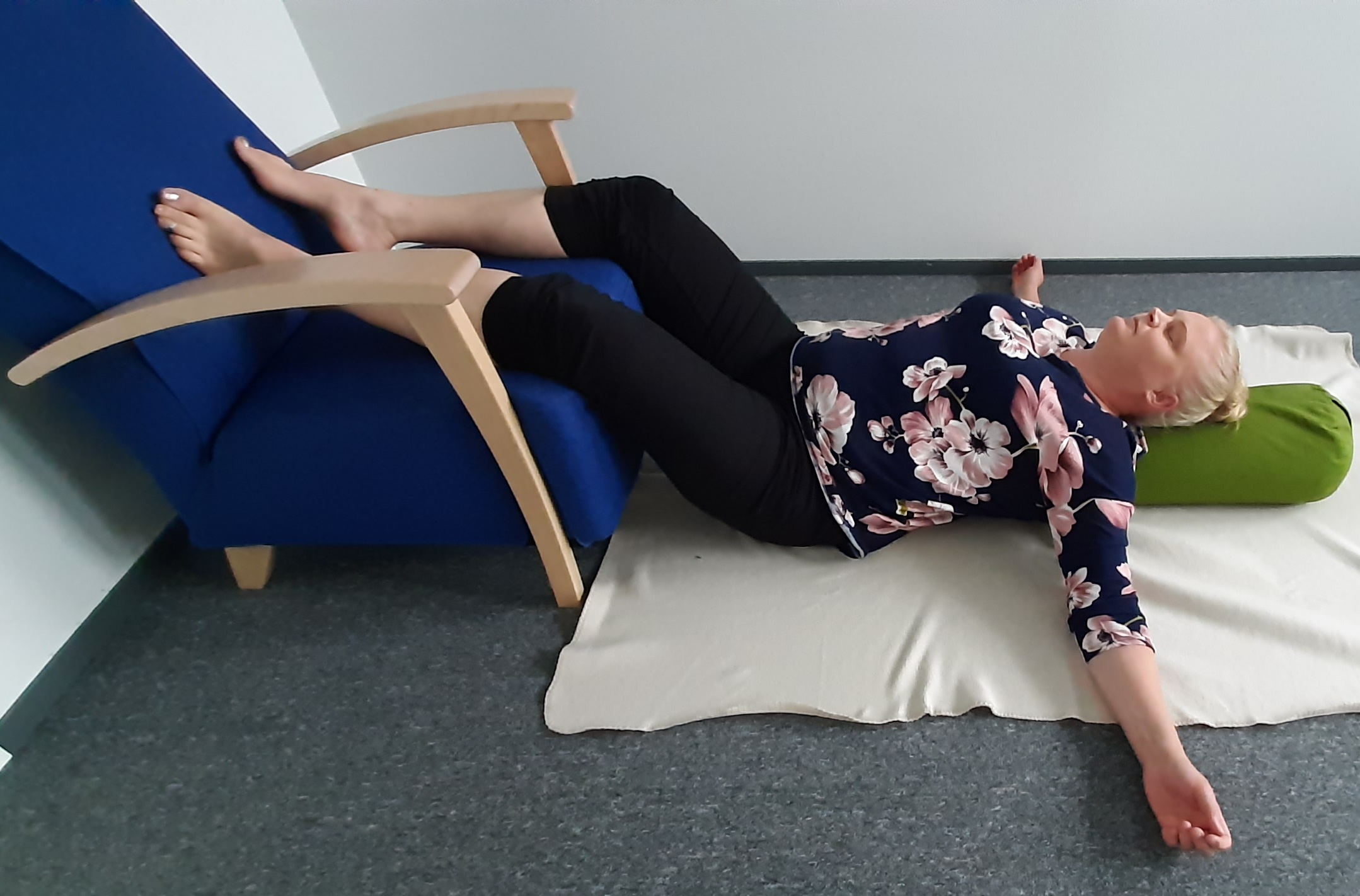 Nainen makaa lattialla selinmakuulla, jalat nostettuina tuolin päälle, kädet vartalon sivuille avattuina. Naisella on joogatyyny selän alla selkärangan suuntaisesti.