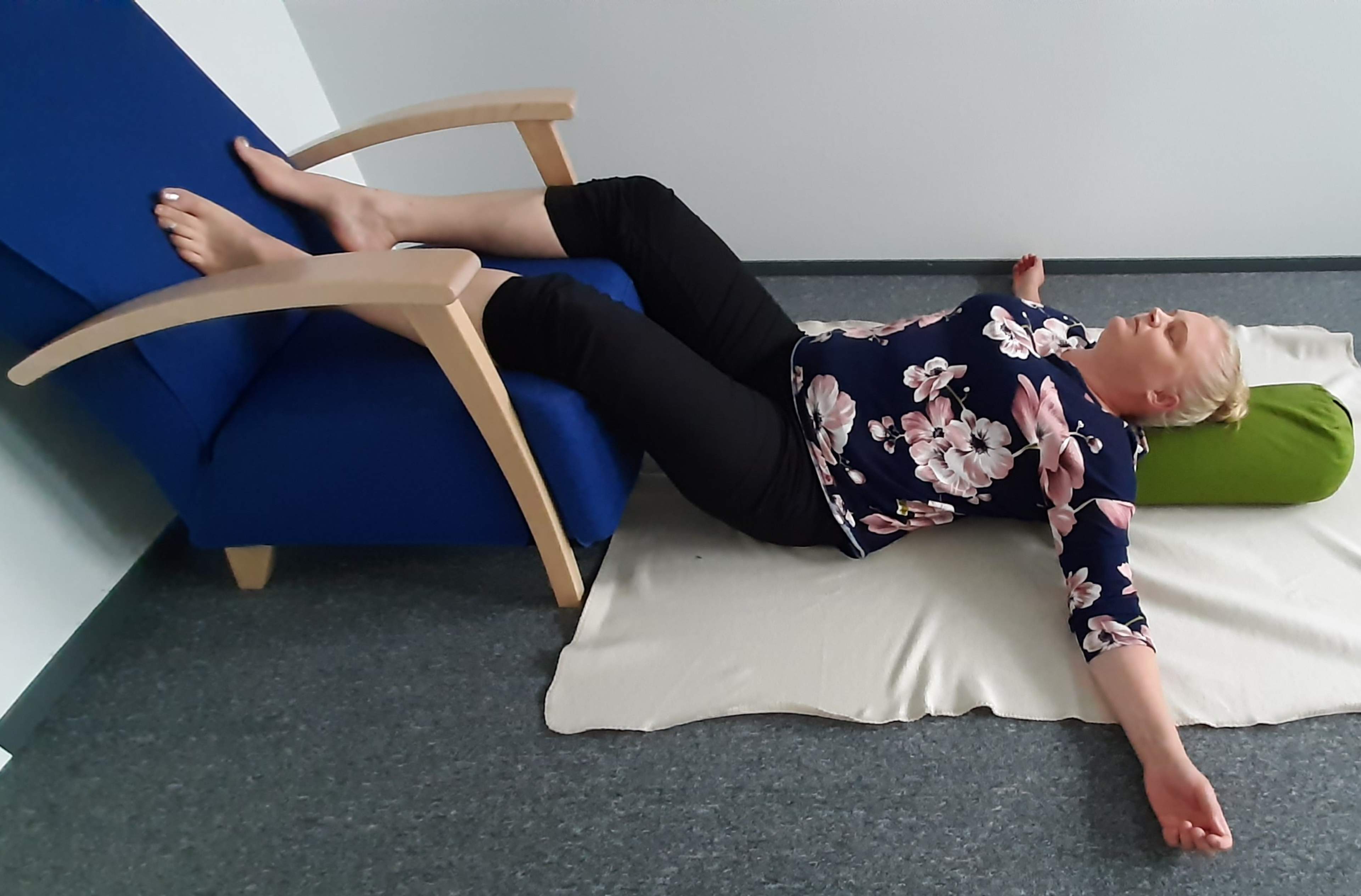 Nainen makaa lattialla selinmakuulla, jalat nostettuina tuolin päälle, kädet vartalon sivuille avattuina. Naisella on joogatyyny selän alla selkärangan suuntaisesti.