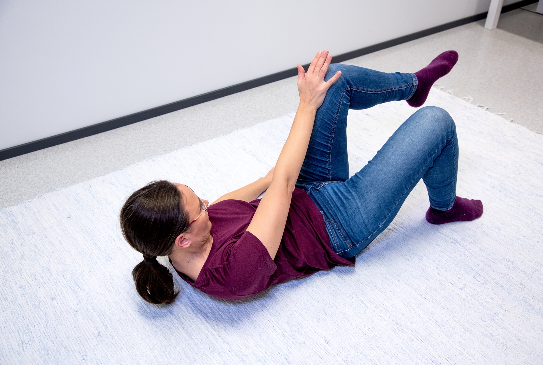 Kvinnan ligger på rygg på golvet med böjda knän och fötterna i golvet. Hon sträcker sig med höger hand mot vänster knä.