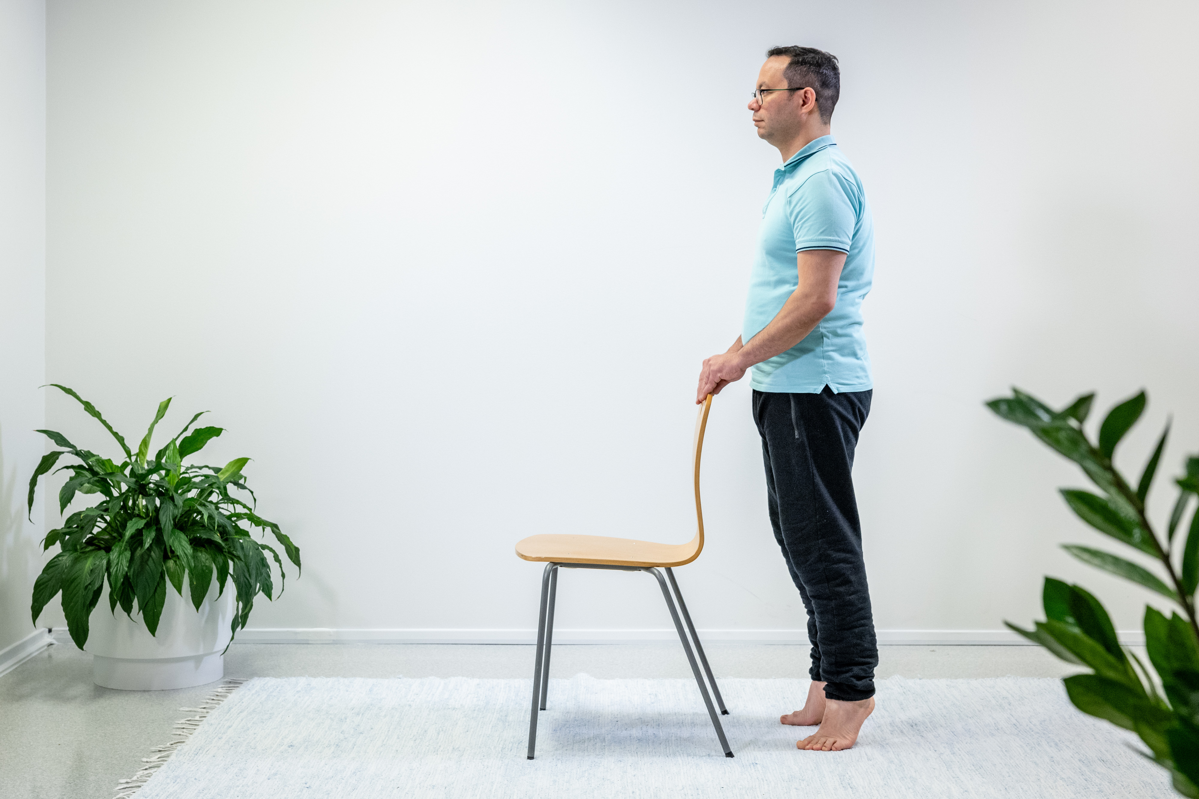 Mies pitää käsillä tukea tuolin selkänojasta ja seisoo varpaillaan. 