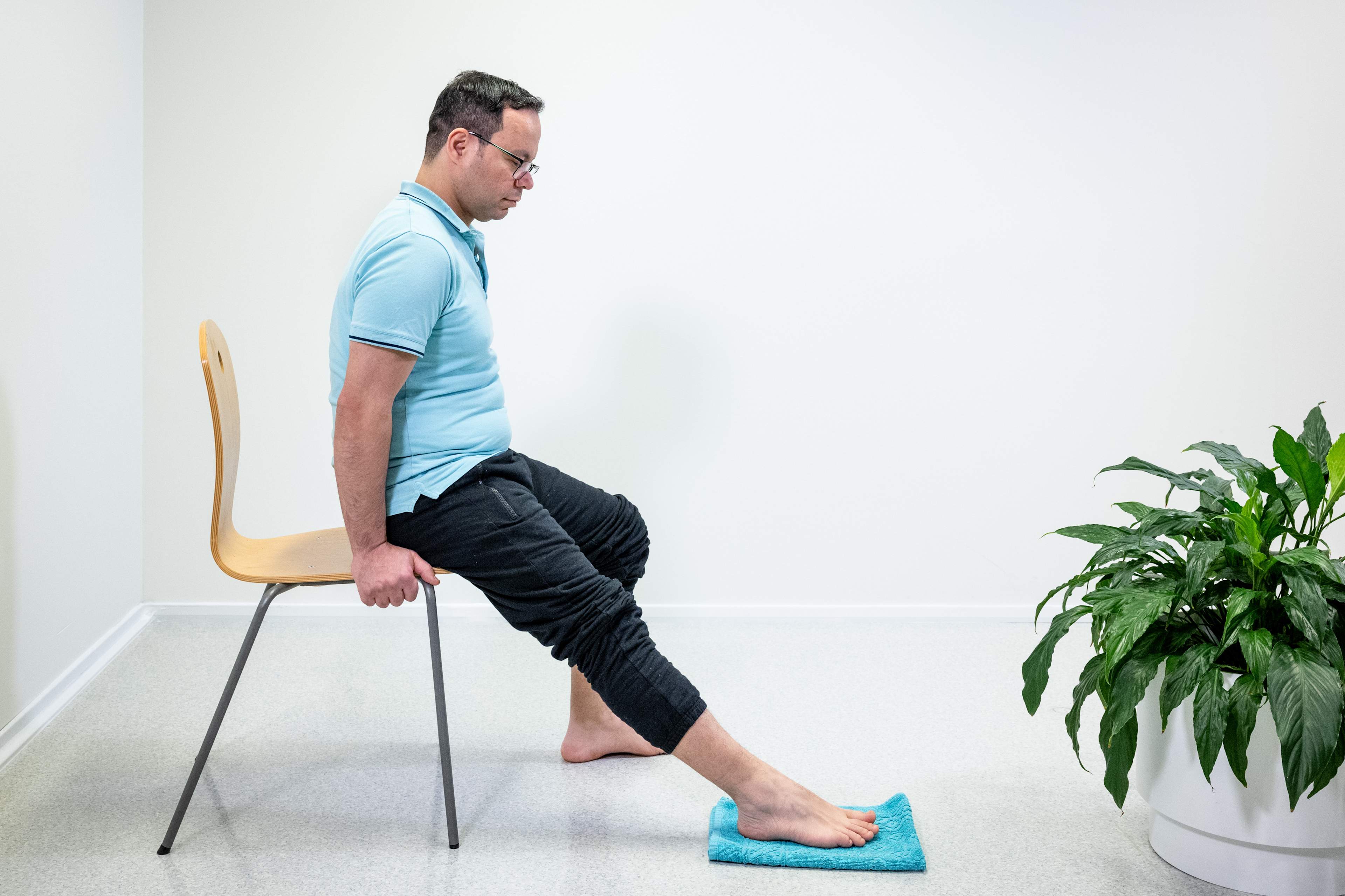 En man sitter på kanten av en stol och har sträckt ut höger ben rakt. Fotsulan är i golvet och det finns en handduk under foten.