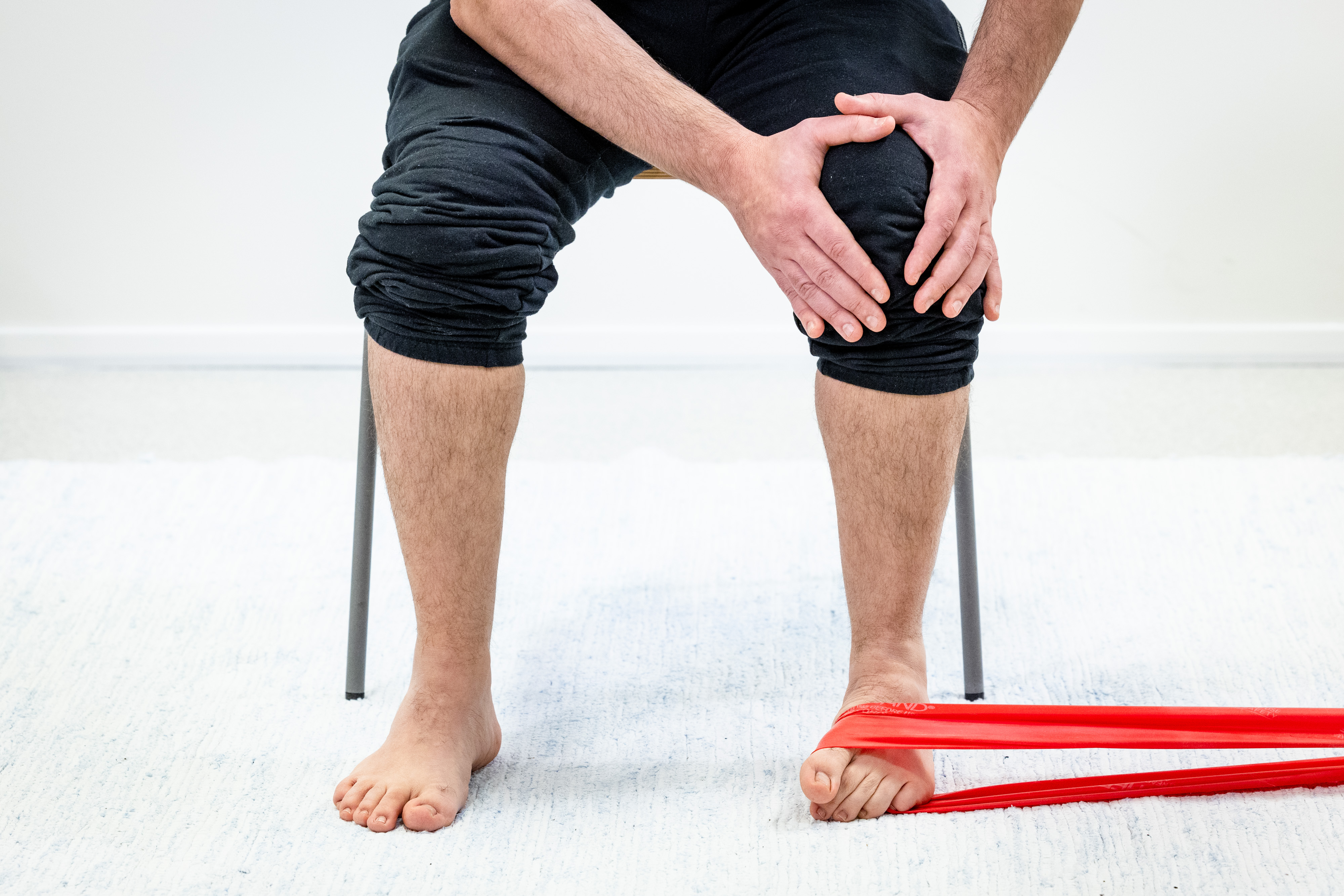 Mies istuu tuolilla ja pitää käsillä vasemmasta polvesta kiinni, vastuskuminauha on vasemman jalkaterän ympärillä ja kiinni esimerkiksi pöydän jalassa. Mies on kääntänyt vasemman jalkaterän isovarpaan suuntaan, kantapää on pysynyt paikallaan.