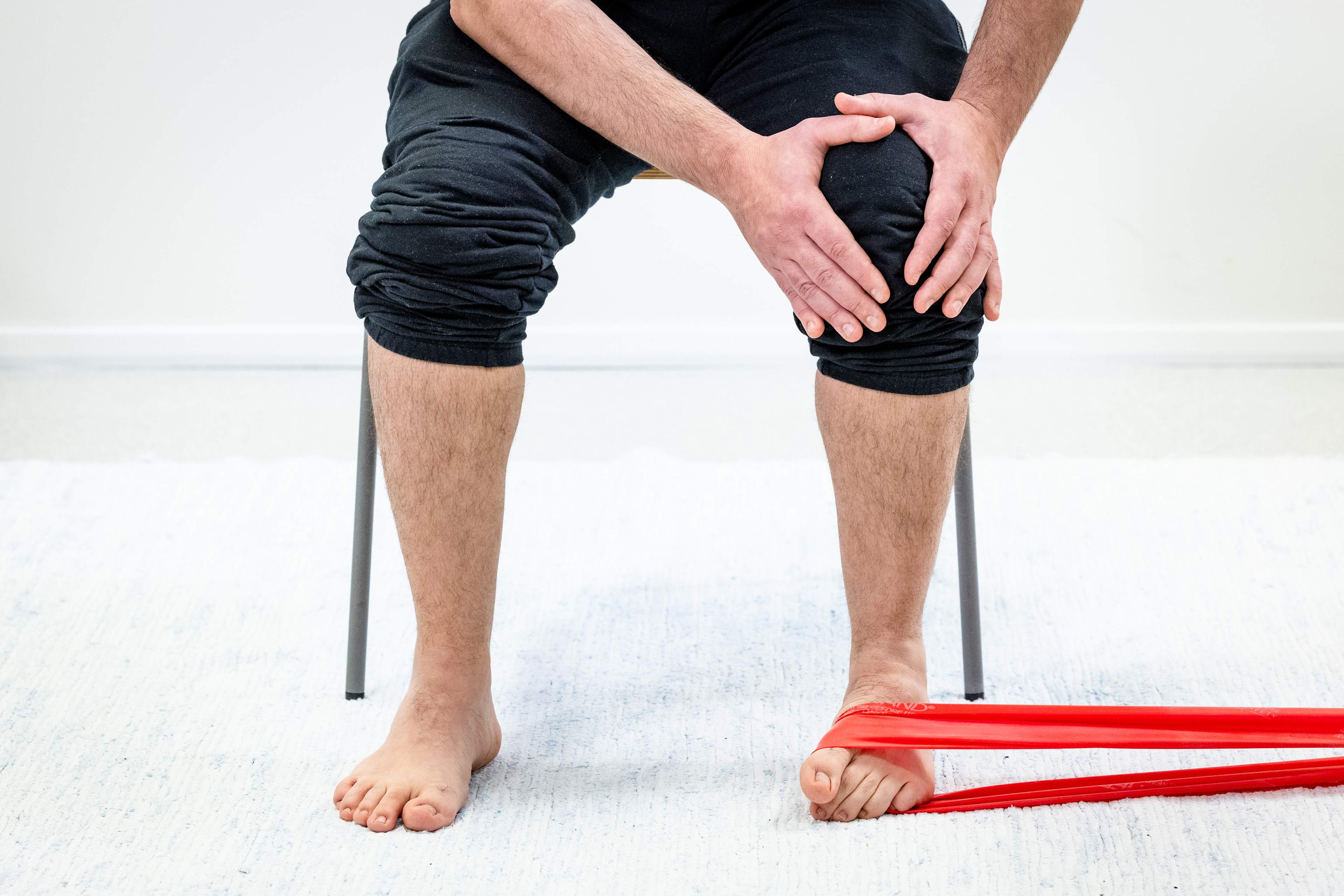 Mies istuu tuolilla ja pitää käsillä vasemmasta polvesta kiinni, vastuskuminauha on vasemman jalkaterän ympärillä ja kiinni esimerkiksi pöydän jalassa. Mies on kääntänyt vasemman jalkaterän isovarpaan suuntaan, kantapää on pysynyt paikallaan.