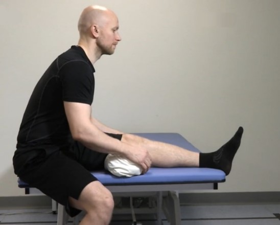 Man sitter på soffkanten, vänster ben upp på soffan och höger ben på golvet. Under vänster knä har mannen en kudde och han lutar sig framåt mot vänster ben.