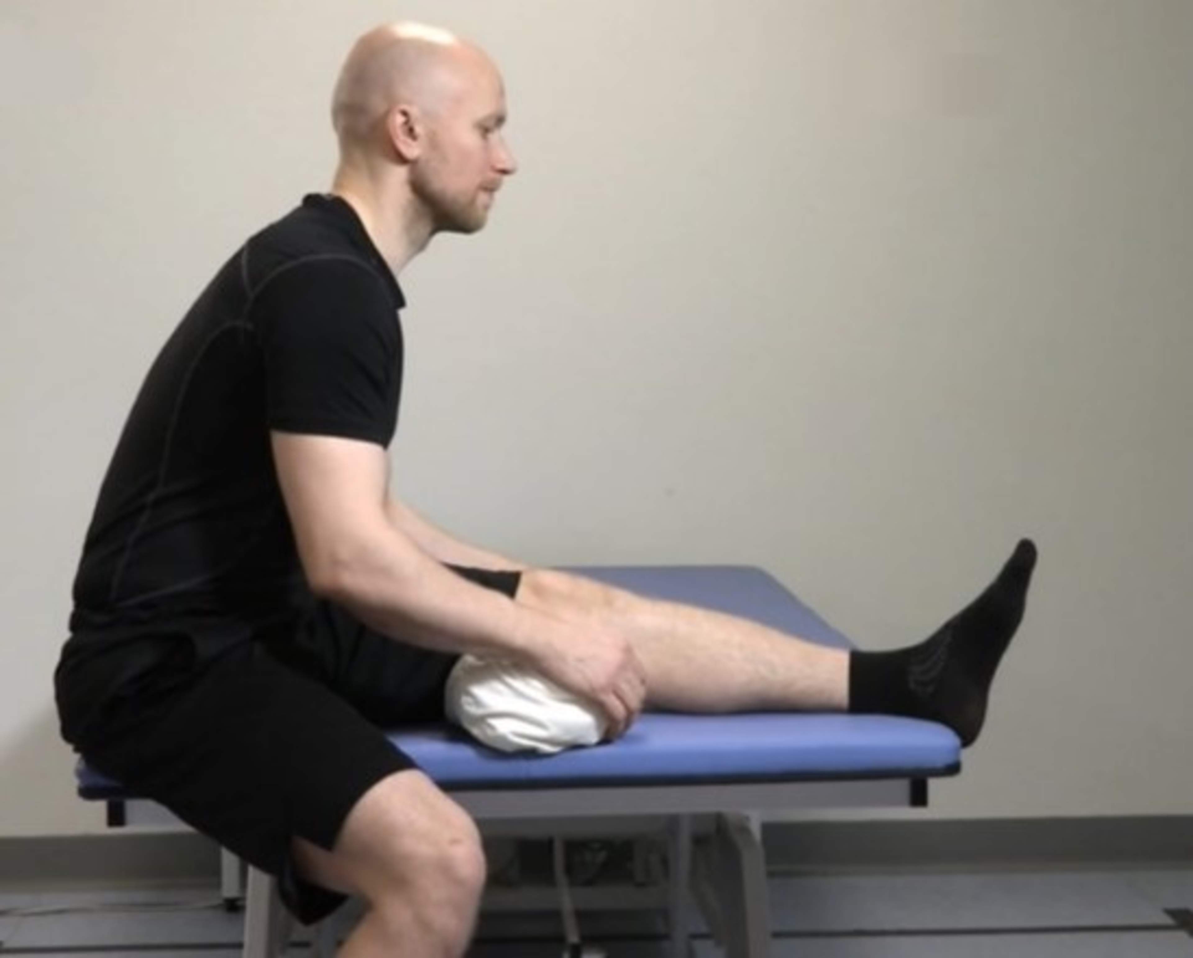 Man sitter på soffkanten, vänster ben upp på soffan och höger ben på golvet. Under vänster knä har mannen en kudde och han lutar sig framåt mot vänster ben.